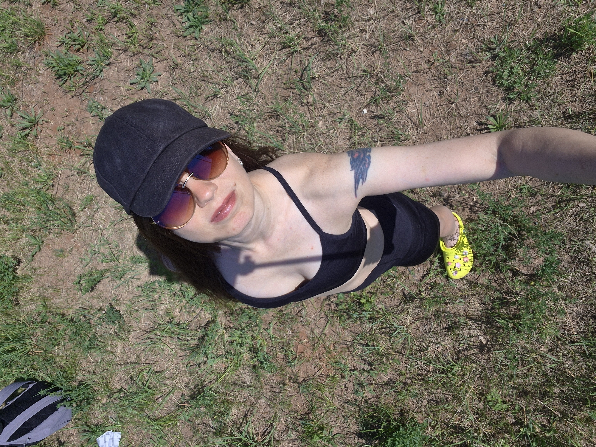 A woman wearing a black cap, sunglasses, and a black top is standing on grassy terrain. She is extending one arm towards the camera, suggesting a selfie. She has a tattoo on her upper arm. She is also wearing a yellow shoe. A backpack is visible in the lower left corner. The location appears to be outdoors, with a mix of grass and bare soil. The exact location in the city of Unknown, Unknown cannot be determined from the image.