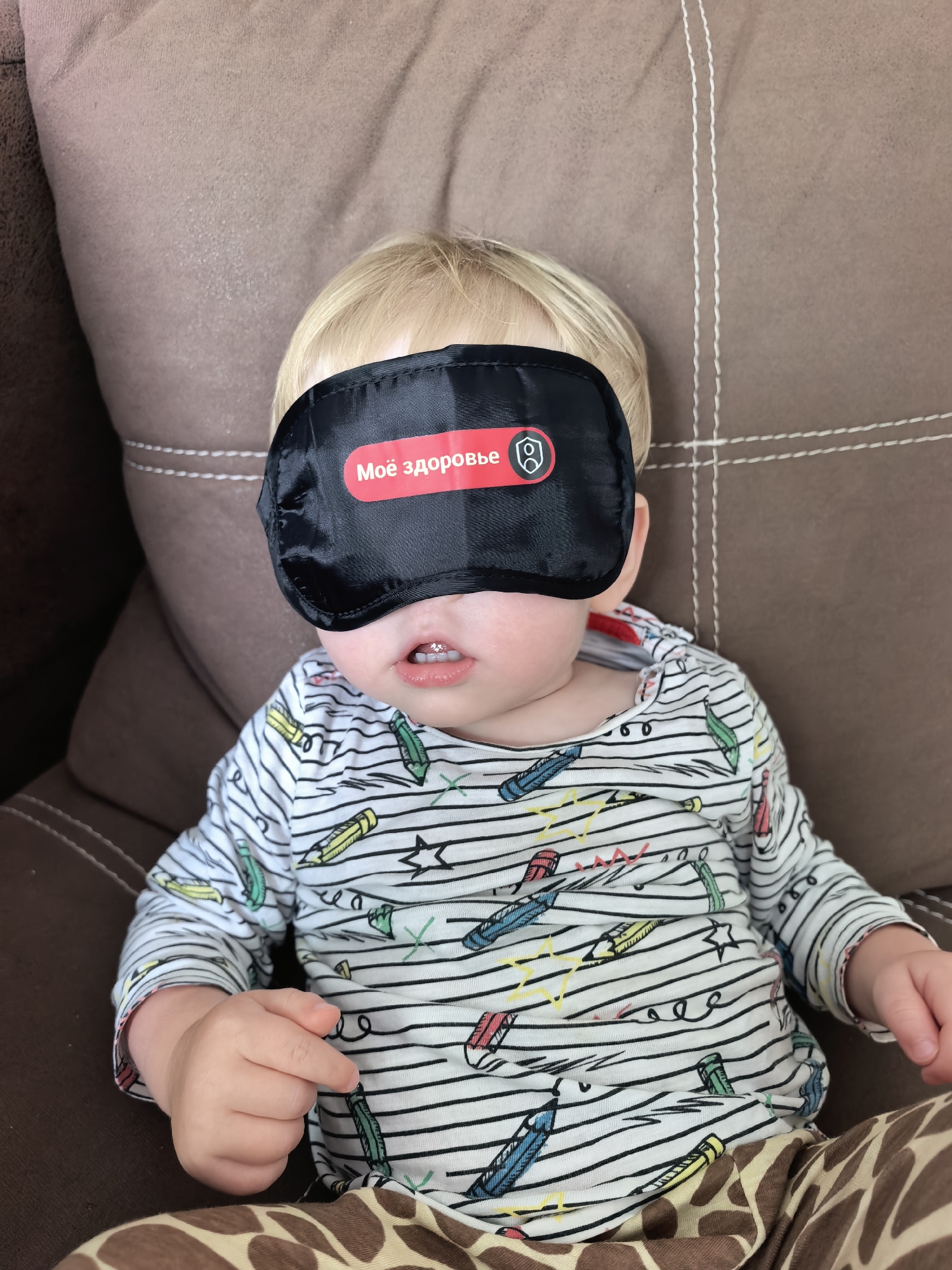 A young child with blond hair wears a black eye mask with red Cyrillic text and a shield icon. The child is lying on a brown couch with visible stitching and has a white long-sleeved shirt with a pattern of pencils and stars. The child's mouth is slightly open, revealing teeth. The image appears to be taken indoors, on a couch. The location of Sterlitamak, Russia is not present in the image.