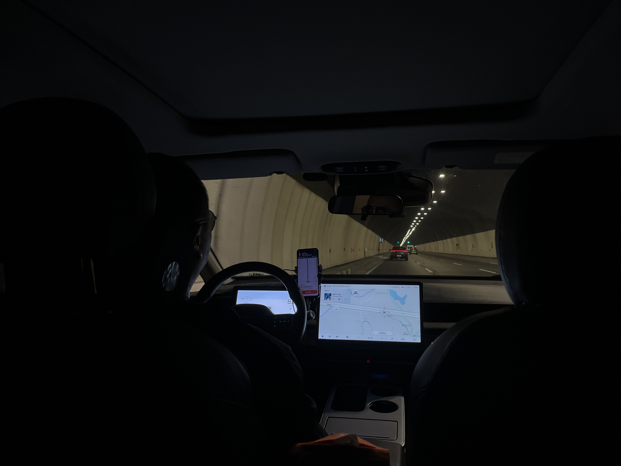 The image shows the interior of a car traveling through a tunnel. The car has a digital display screen showing a map interface and a phone mounted near the steering wheel with a navigation application running. Through the windshield, the tunnel interior, other vehicles, and tunnel lighting are visible. The photograph is taken from the back seat of the vehicle looking forward, with the dark silhouettes of the front seat passengers. The GPS coordinates on the main display screen indicates the location is in the Chongqing High-tech Zone, China.