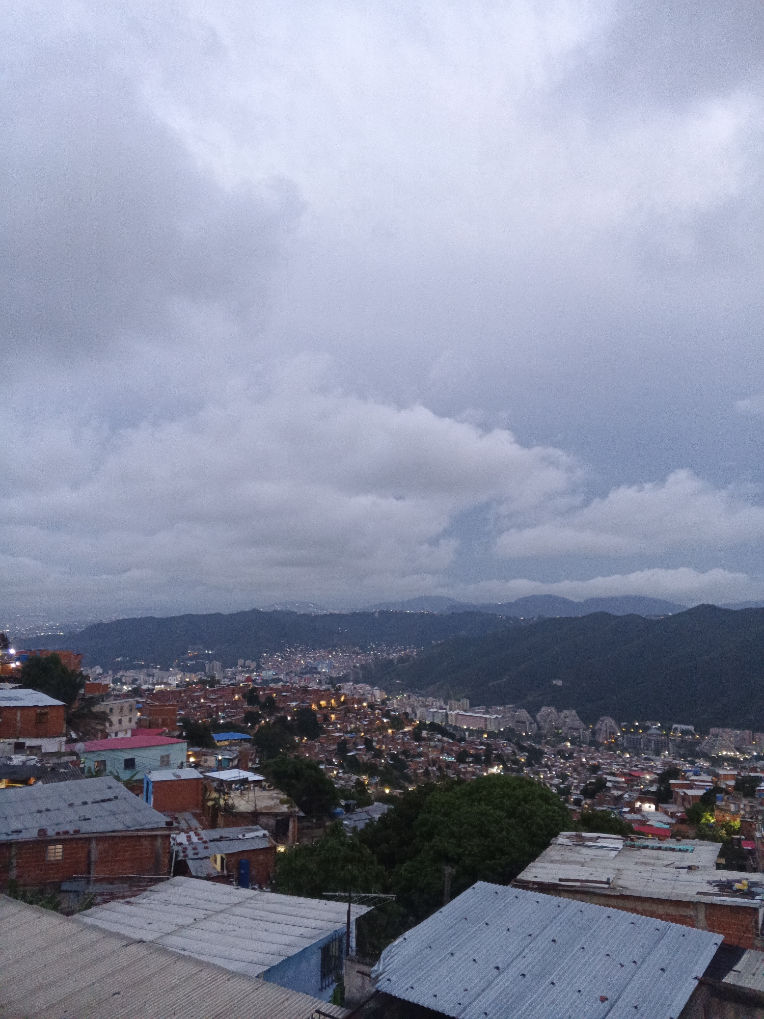 The image is a photograph taken from a high vantage point, likely a hillside, looking out over a sprawling city at dusk. In the foreground, there are several rooftops made of corrugated metal and what appears to be brick structures, suggesting a residential area, possibly a barrio or favela. The houses are densely packed together and vary in color and material.
Beyond the immediate foreground, the city stretches out into the distance. Numerous buildings and lights are visible, indicating an urban environment. The city is nestled in a valley with dark, tree-covered mountains in the middle ground and background. The sky is overcast with heavy, gray clouds, and the overall lighting suggests either early morning or late evening.
The scene is characteristic of the hilly topography of Caracas, Venezuela, where many neighborhoods are built on steep slopes. The presence of a dense, informal settlement in the foreground, overlooking a more developed urban area in the distance, is also typical of the city's socio-economic landscape. The photograph captures a sense of the vastness of the urban sprawl and its relationship with the surrounding natural environment. There are no people or specific actions depicted in the image; it is a still, environmental portrait of the city.