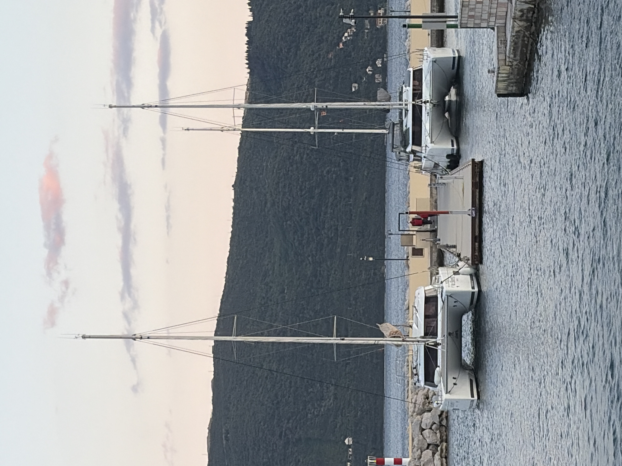 This image captures a serene marina scene in Meljine, Montenegro, likely during late afternoon or early evening, given the soft, muted lighting and pastel hues in the sky.
The primary subjects are two modern, white sailboats, appearing to be catamarans, docked side-by-side at a pier. Both boats feature tall, slender masts with intricate rigging reaching towards the sky. The closer boat is partially obscured by a rocky shoreline or breakwater in the foreground.
Behind the marina, a prominent, dark green, forested mountain rises, filling the mid-ground. Scattered across the lower slopes of this mountain, small buildings and houses are subtly visible, blending into the natural landscape.
The water in the marina is calm with gentle ripples, reflecting the ambient light. A concrete or stone dock runs parallel to the boats, and a darker, wooden-looking section of the pier extends into the water. On this dock, near the foreground, a small, vertical sign is visible, with faint, almost illegible text that might read "GAFE 7" or a similar berth identifier. No people are visible, contributing to a peaceful and tranquil atmosphere. The sky above is largely clear but features elongated, wispy clouds brushed with faint pink and purple tones, characteristic of twilight.