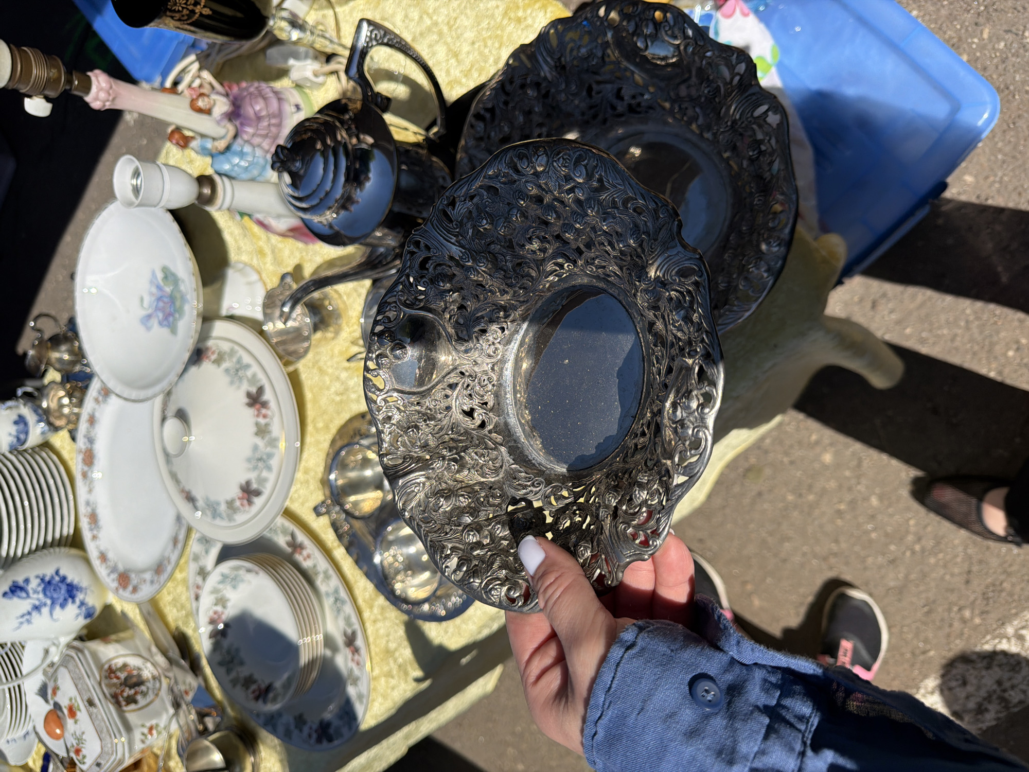 This image captures a close-up, high-angle view of an outdoor market stall in Riga, Latvia, under bright, direct sunlight. The stall's table is covered with a yellow, textured fabric. A diverse array of antique or vintage items is densely arranged on the table.
**Objects on the Table:**
The collection includes numerous ceramic and porcelain pieces:
* Multiple stacks of white plates, some adorned with floral patterns in blue, red, green, and gold.
* A prominent white tureen or covered serving dish with elaborate floral and figural motifs in orange, brown, and green.
* Several bowls, including small ones with pink and green floral designs and a footed bowl with light blue floral patterns.
* A white pitcher featuring blue floral designs.
* An oval platter and a covered dish, both with light blue, pink, and green floral patterns.
* Silver or silver-plated items include a fluted teapot, several small ornate dishes with intricate openwork scroll and floral designs, and possibly a creamer and sugar bowl set.
* A ceramic lamp base depicting two women in period dresses with a visible light socket above, and a simpler white and brown lamp base.
* A black, ornate drinking glass or vase with gold detailing.
**People and Actions:**
In the foreground, a person's left hand and forearm are visible. The hand, with light skin and white nail polish on the thumb, index, and middle fingers, is holding one of the round, ornate silver-plated dishes, appearing to examine or present it. The person wears a blue long-sleeved shirt with a visible dark blue button on the cuff. A black shoe with pink accents is partially visible on the ground to the lower left, and another person's black mesh shoe is visible further right.
**Environment:**
The scene is brightly lit by direct sunlight, casting distinct shadows from the displayed items and the person's hand onto the table and ground. The ground beneath the table and feet is concrete or asphalt. A blue plastic storage container is partially visible under the table on the right side. The background is largely blurred, suggesting an open outdoor setting.