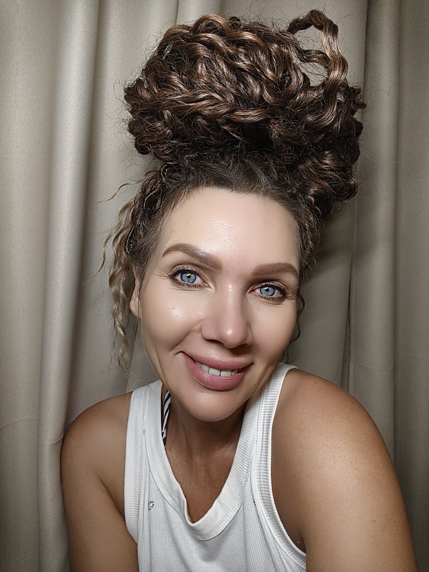 This is a medium shot, head and shoulders portrait of a woman with a wide, genuine smile. She has striking blue eyes and light skin with a healthy glow. Her brown, curly hair is styled in a large, voluminous bun atop her head, with a few tendrils framing her face. She is wearing a white ribbed tank top, revealing a glimpse of a striped pattern underneath. The background consists of soft, beige curtains, suggesting an indoor setting. The lighting appears to be soft and diffused, possibly from an artificial source, creating a warm and inviting atmosphere. The woman's expression conveys happiness and confidence. There is no visible text in the image. The time of day and weather are not discernible from the image.