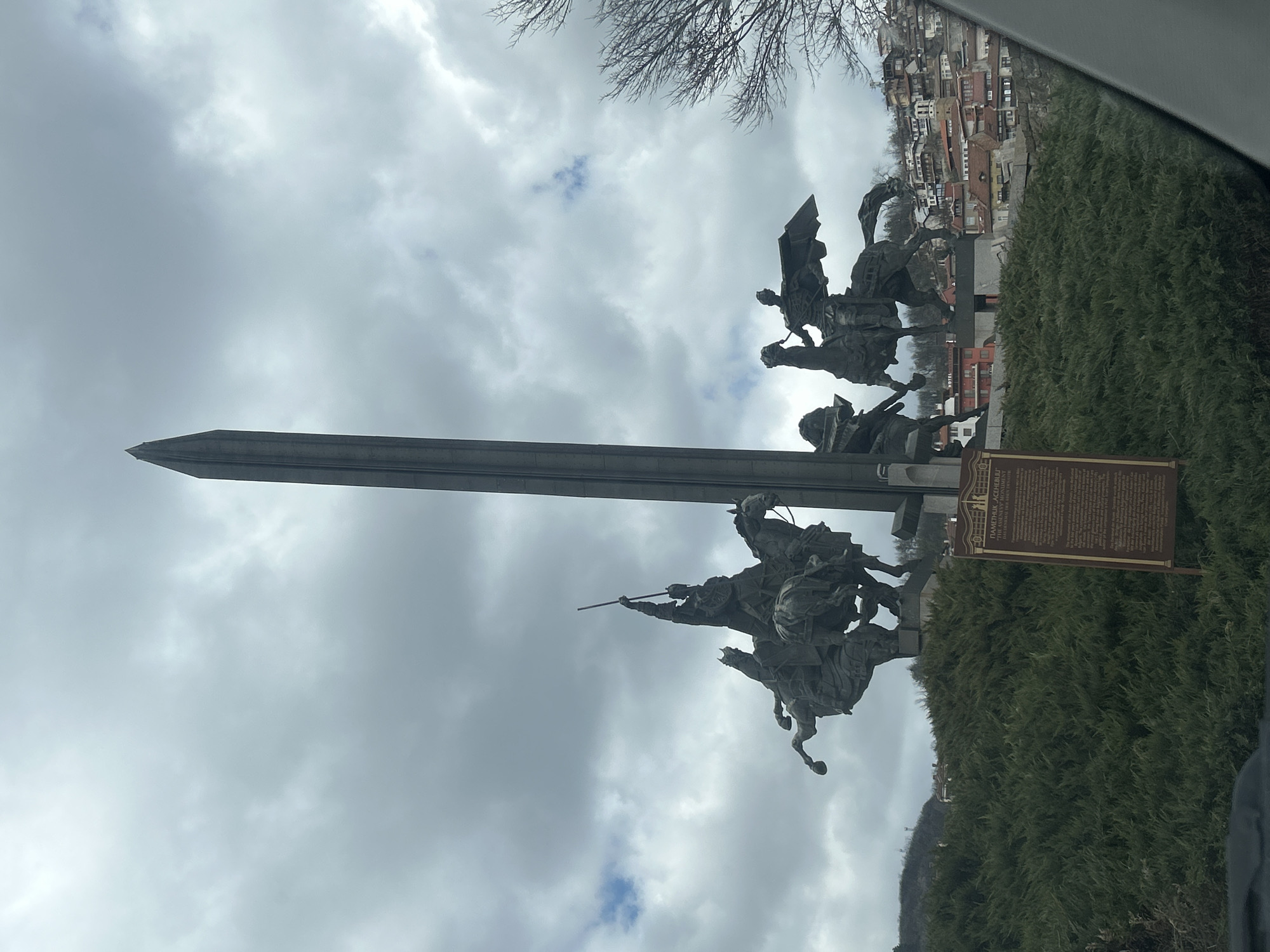 The image depicts the Asenevtsi Monument in Veliko Tarnovo, Bulgaria, set against a cloudy sky. The monument features four bronze figures of Tsars Asen, Peter, Kaloyan, and Ivan Asen II, mounted on horses. A large sword-shaped obelisk rises behind the equestrian figures. A sign with text is located to the right of the statues. In the background, buildings are visible, situated on a hillside. The monument is surrounded by greenery.