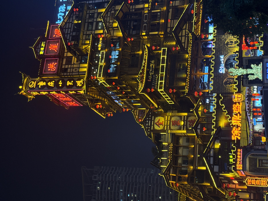 The image displays the multi-tiered Hongyadong Commercial Street complex at night, located in Yuzhong District, Chongqing, China. The scene is dominated by intricate traditional Chinese architecture, characterized by cascading wooden structures, tiled roofs, and numerous balconies, all extensively illuminated with warm yellow lights outlining eaves and windows. Numerous red spherical lanterns hang from various levels. Prominent red neon signs feature Chinese characters, including "洪崖洞" (Hongyadong) on an upper section, and "巴渝小吃城" (Bayu Snack City) on a vertical tower-like element. Other signs for businesses such as "老灶火锅" (Laozao Hotpot) and "涂鼎盛" (Tu Dingsheng) are visible on lower tiers. A large golden character "金" (Jin) is displayed on a mid-level facade. A large arched stone-like gateway is visible near the bottom center, in front of which stands a glowing, light-green illuminated statue. A tall, modern, grey high-rise building with some illuminated windows is visible in the dark background to the left, contrasting with the traditional complex. No individuals or specific actions are discernible. The environment is an urban setting at night.