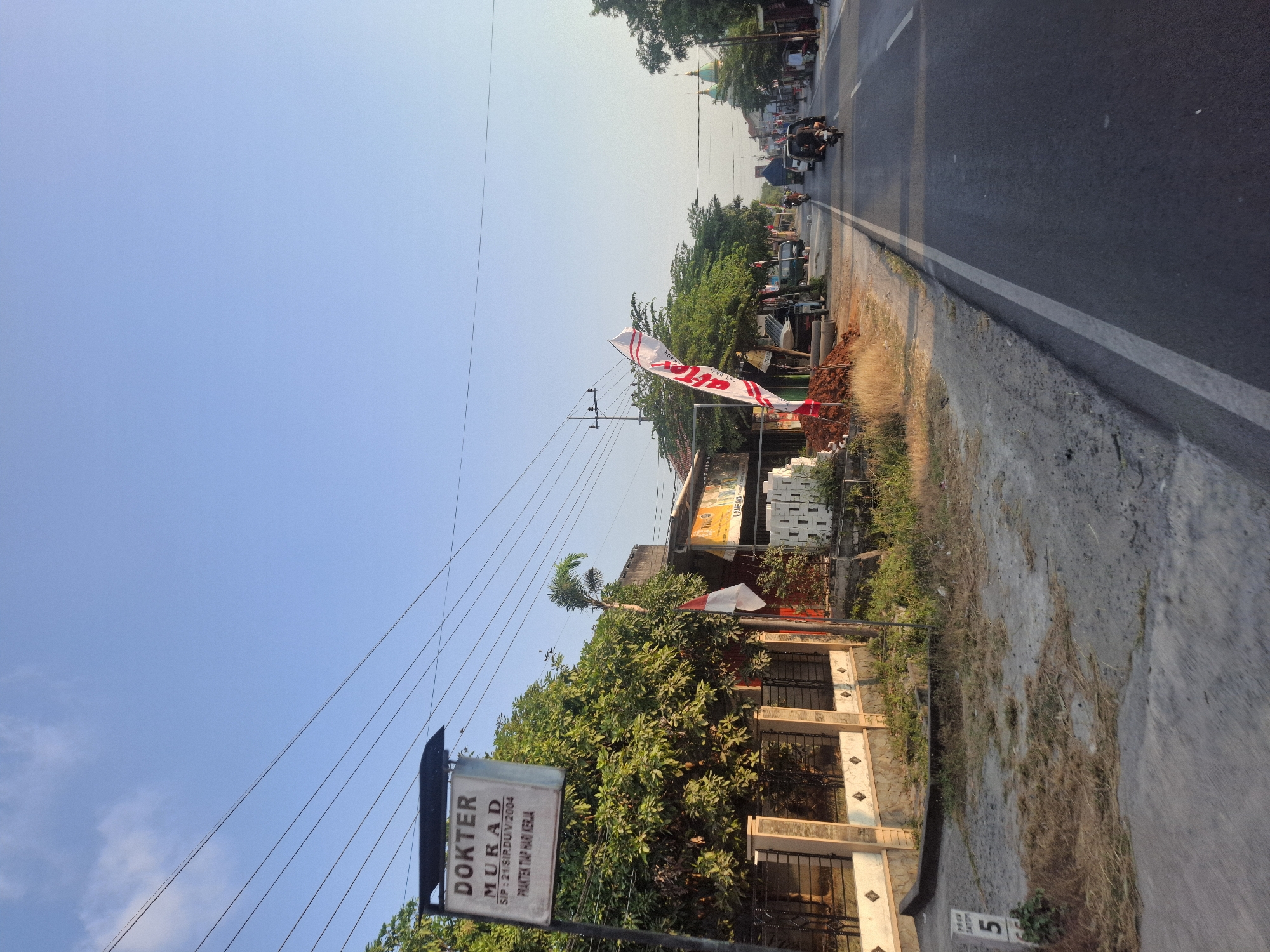 The image captures a roadside scene in Sumberwudi, Indonesia, on a clear day with a bright blue sky. The foreground shows a paved road with a white line marking the lane. To the left of the road is a dirt and gravel area with dry grass and some vegetation. A white sign with the number "5" is visible on the left side of the road. Further back on the left, a fence with decorative stone and metalwork separates the road from a property with lush green trees. A prominent sign for "DOKTER MURAD" is mounted on a pole in front of the trees, with additional text "SIP: 21/SIRDU/V/2004 PRAKTEK TIAP HARI KERJA" (Practice Every Working Day). A small Indonesian flag is displayed on a pole near the sign.
Buildings line the right side of the road, with awnings and structures suggesting commercial or residential use. Several white bricks are stacked in front of one building. A large white banner with red lettering, featuring a stylized flame or wave design, is planted in the ground near the road. Traffic is present on the road, including several motorcycles with riders, a car, and a truck. The overall impression is of a moderately busy street in a semi-urban or rural setting. The presence of the doctor's sign and the style of the buildings suggest a local community setting.