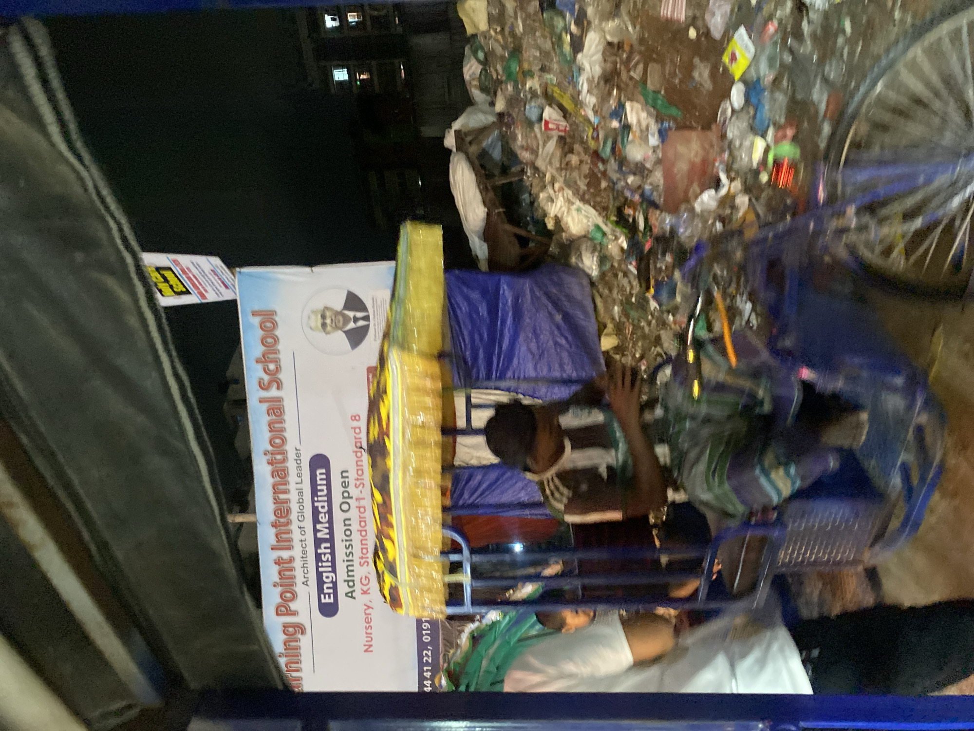 The image captures a nighttime scene in Dhaka, Bangladesh, with a clear contrast between a school advertisement and a pile of garbage. A prominent banner for "Learning Point International School" is visible, advertising "English Medium" education and "Admission Open" for Nursery, KG, Standard 1-Standard 8. A circular portrait of a man in a suit is on the banner. To the right of the banner, a large accumulation of trash, including plastic bottles, bags, and assorted debris, covers the ground. In the foreground, a blue rickshaw is partially visible. A person wearing a striped shirt is seated in the rickshaw, appearing to be operating it or engaged in some activity within it. Another person, wearing a white shirt, is standing to the left of the rickshaw. The overall environment appears to be an urban setting, likely on a street or open area, given the presence of the rickshaw and the buildings in the background, which show illuminated windows. The lighting suggests it is nighttime, with artificial light sources illuminating the scene.
