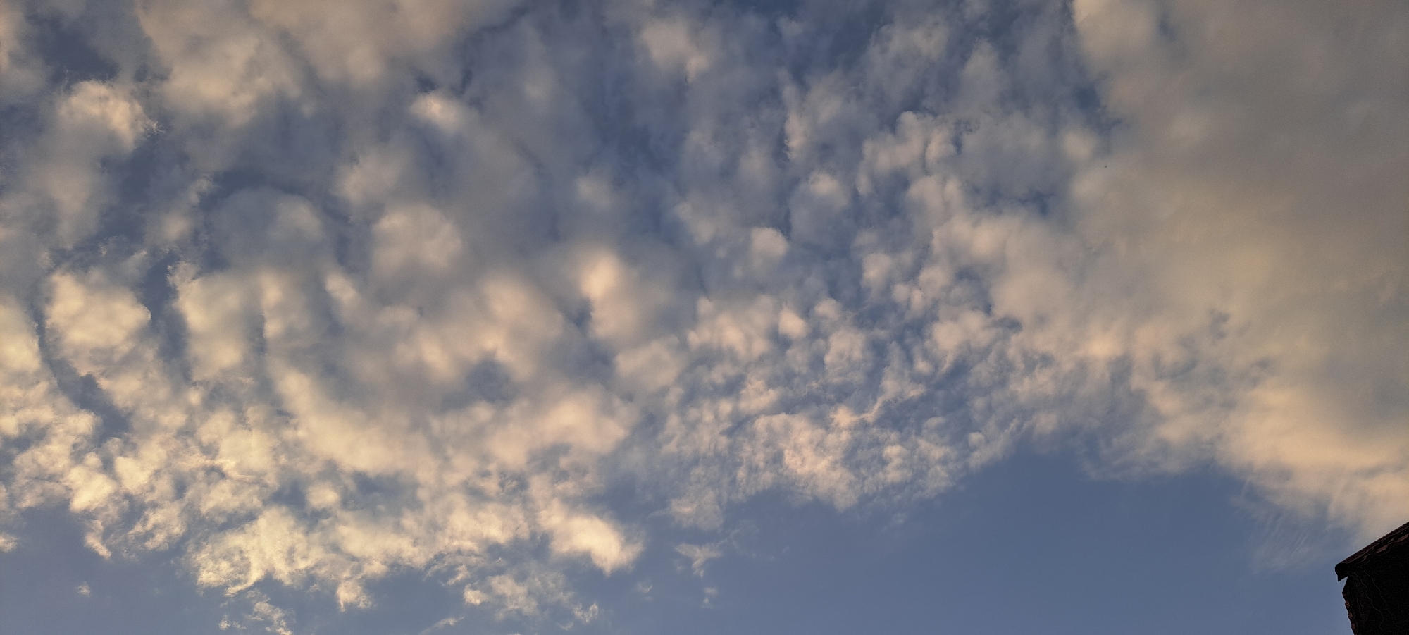 The image depicts a sky filled with scattered clouds, illuminated by a warm, possibly sunset or sunrise light, casting a soft glow on their undersides. The sky is a clear blue, with the clouds varying in density and texture, ranging from wispy streaks to more substantial, puffy formations. In the lower right corner of the frame, a dark, angled roofline of a building is visible, suggesting an outdoor, possibly urban or semi-urban, setting. The overall impression is one of a serene sky, possibly observed from a vantage point within or near the city of Bash-Kaiyndy, Kyrgyzstan. No people or animals are visible in the frame.
