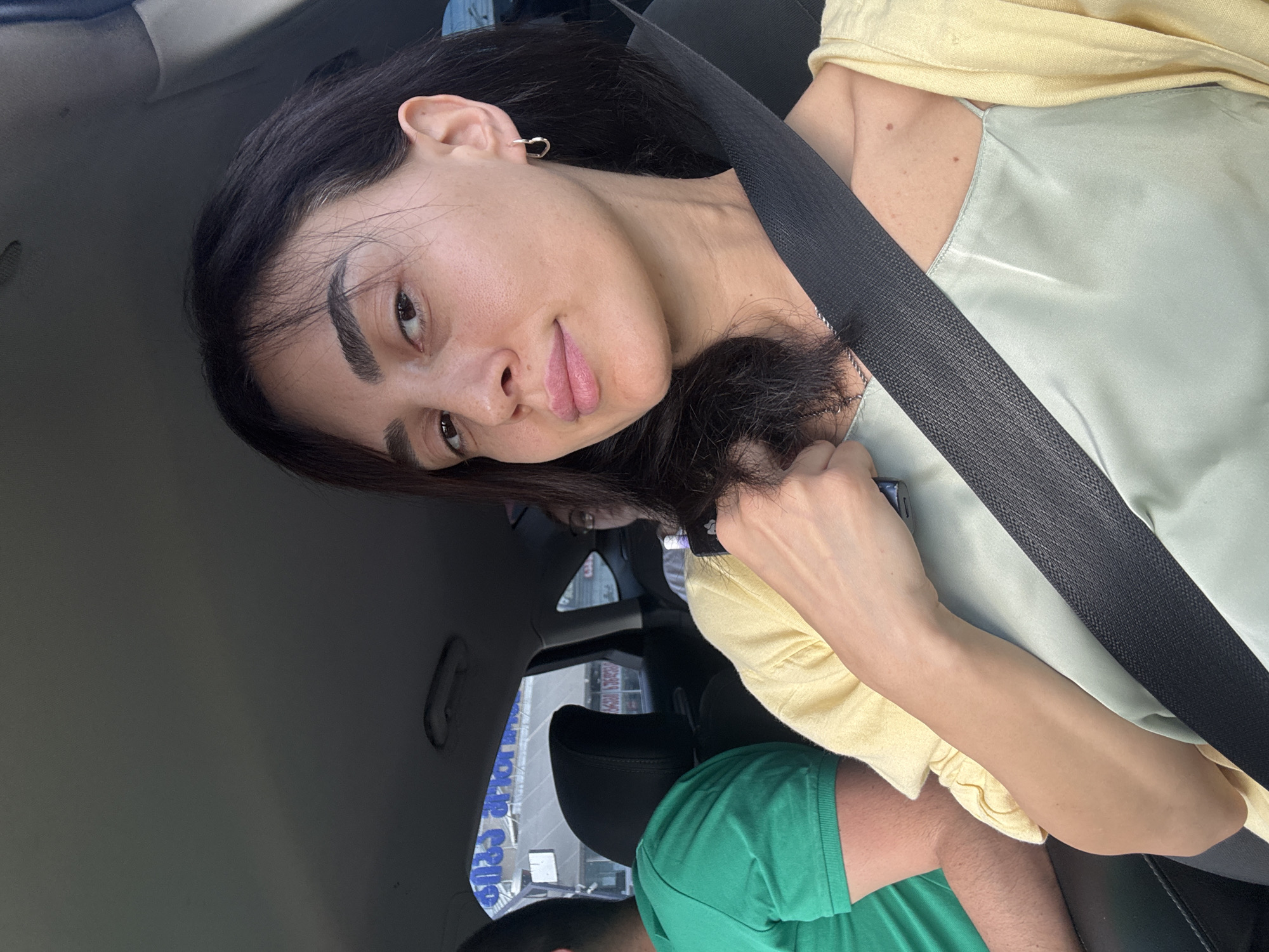 A woman is sitting in the passenger seat of a car, wearing a yellow cardigan over a light green slip dress and a seatbelt. She has dark hair, fair skin, and is wearing a small hoop earring in her right ear. She is holding a dark object in her right hand. A man in a green shirt is visible in the driver's seat, and another person with glasses is in the back seat. The car appears to be driving through the city of Tbilisi, Georgia, as indicated by the text on the building visible through the car window, which reads "საქართველოს ბანკი" (Georgian Bank). The building is modern and appears to be a commercial establishment. The overall scene depicts a casual moment of travel within the city.
