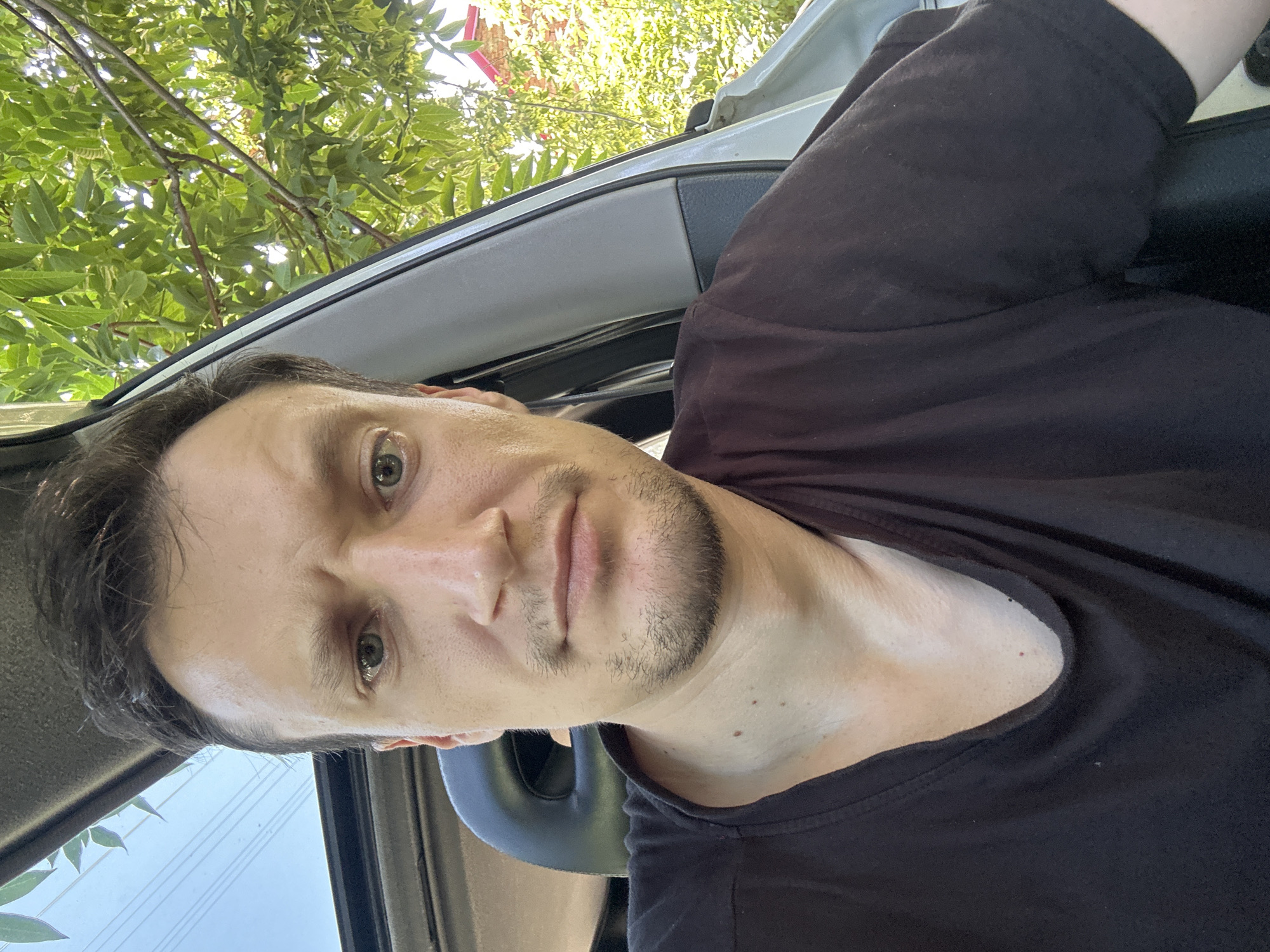 A close-up, selfie-style photograph captures a man from the chest up, seated inside a vehicle. He is wearing a plain black t-shirt. The man has medium-length, dark brown hair and a light beard. His eyes are open and he is looking directly at the camera with a neutral expression. Sunlight filters through the car's sunroof and the leaves of a tree located outside the open car door, casting dappled light on his face and the surrounding interior. The background is dominated by the green foliage of the tree and hints of a brick building visible through the leaves. The car's interior shows a dark seat and the edge of the door frame. The image is taken during daylight hours, likely outdoors in a rural or suburban setting. Based on the provided context, this scene is located in Makiv, Ukraine.