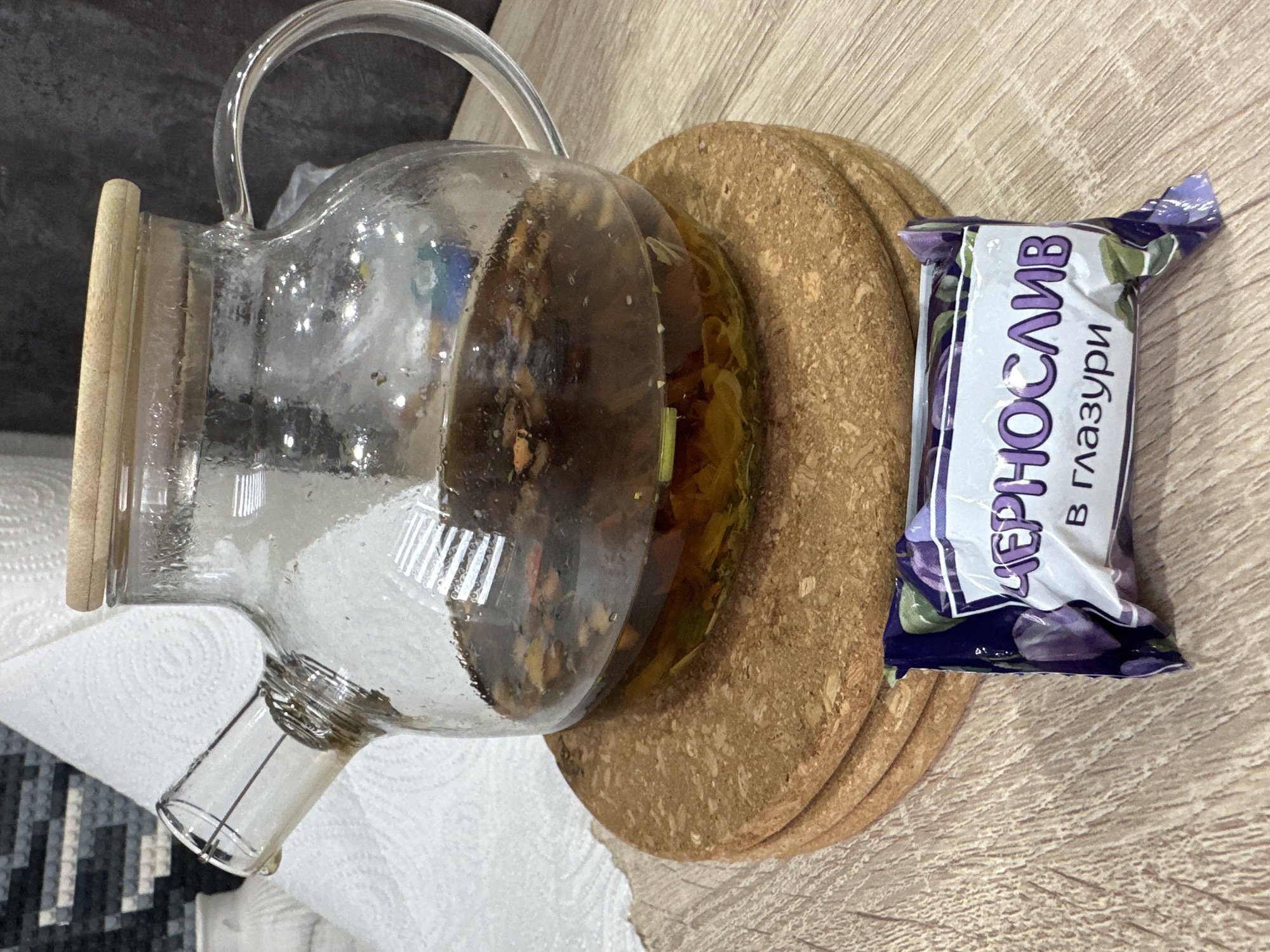 A glass teapot filled with a dark liquid and what appears to be dried fruit or herbs sits on a stack of three cork coasters. In front of the teapot, a wrapped candy with purple fruit imagery and Russian text that translates to "Prune in glaze" is placed on a light-colored wooden surface. In the background, a white textured surface, possibly paper towels, is visible, along with a dark, textured wall. The lighting is soft and casts subtle reflections on the glass. The scene appears to be indoors, likely in a kitchen or dining area. There is no indication of the specific location within Chaikovsky, Russia.