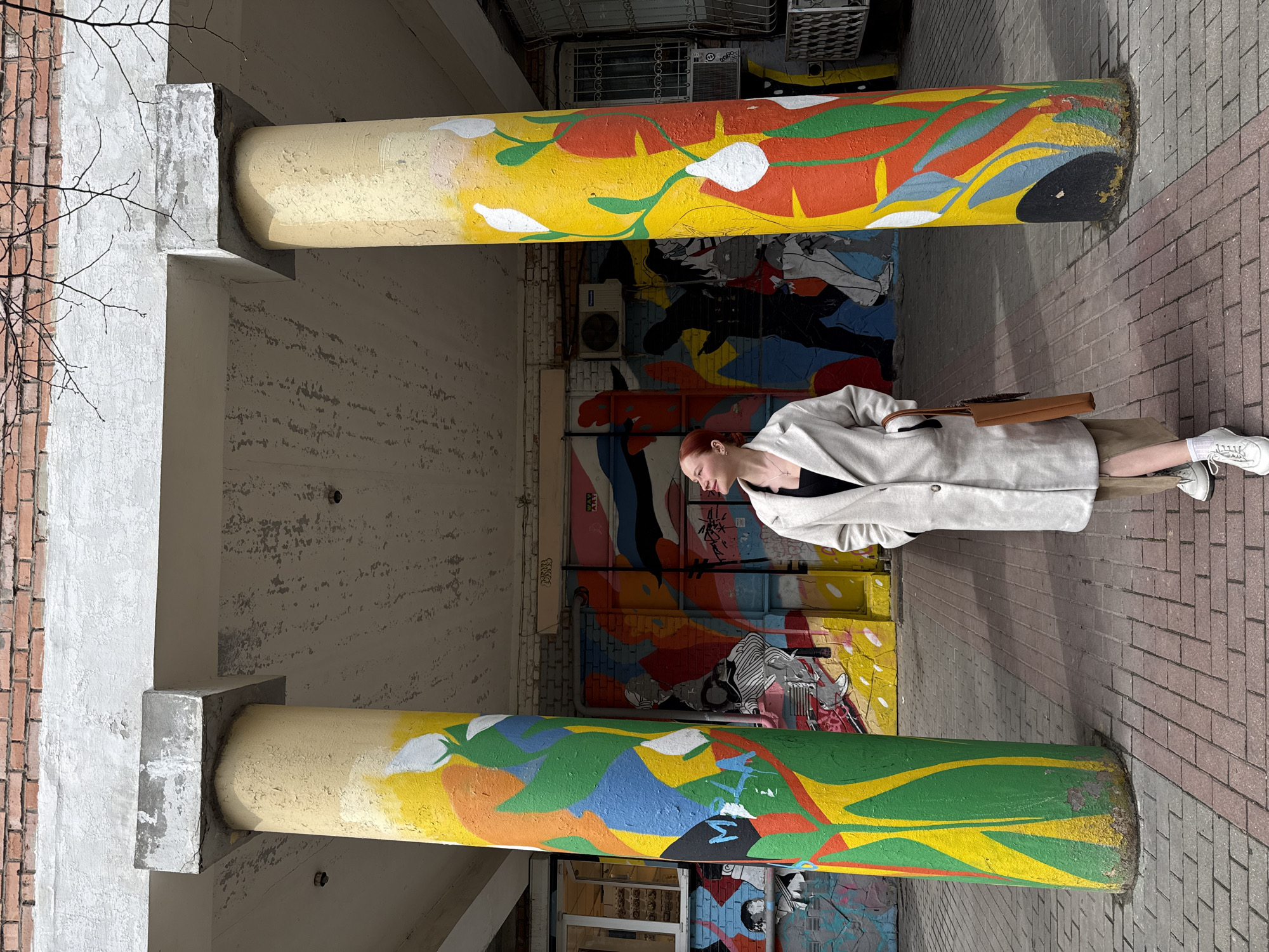 The image shows a woman standing on a brick walkway between two painted columns. The columns are part of a building's support structure. The walls behind the columns are covered in graffiti-style artwork. The woman is wearing a light-colored coat, a dark top, a tan skirt, white socks and white shoes, and carrying a brown purse. The overall location is characterized by colorful graffiti art and urban architecture. This appears to be Ovoshchnoy, Russia, based on the style of building construction and graffiti.