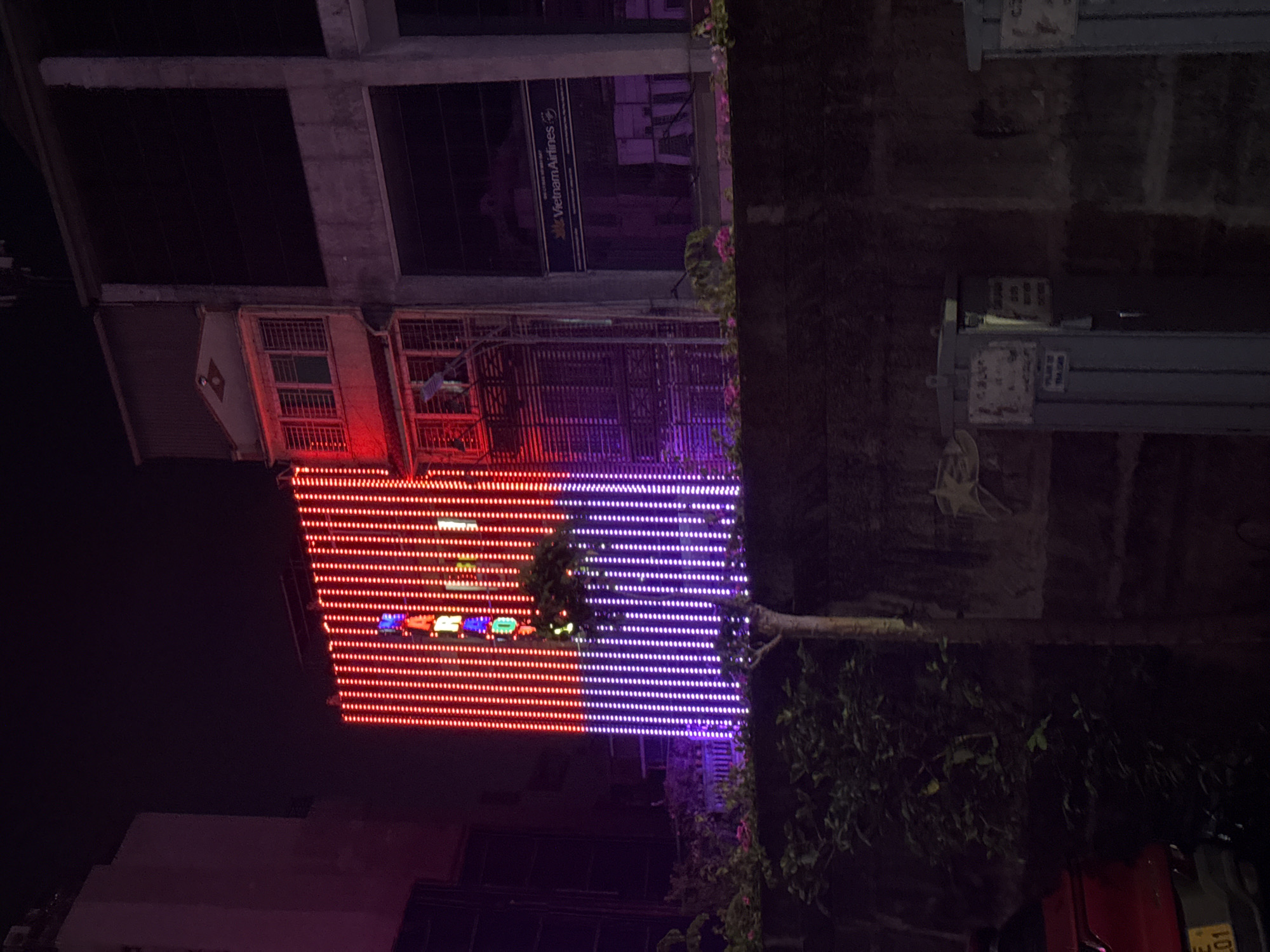 The image captures a nighttime street scene in Hanoi, Vietnam, dominated by a building facade adorned with a vertical display of red and purple LED lights. The lights form a pattern that is partially obscured by a small tree and surrounding vegetation in the foreground. To the right of the LED display, a section of a building is visible, featuring a sign for "Vietnam Airlines." The image also shows a dark, textured wall in the lower portion, with some visible graffiti. A red car is partially visible on the left side of the frame, with a portion of its license plate showing "01". The overall atmosphere is urban and nocturnal, with the bright lights of the building creating a stark contrast against the dark night sky.