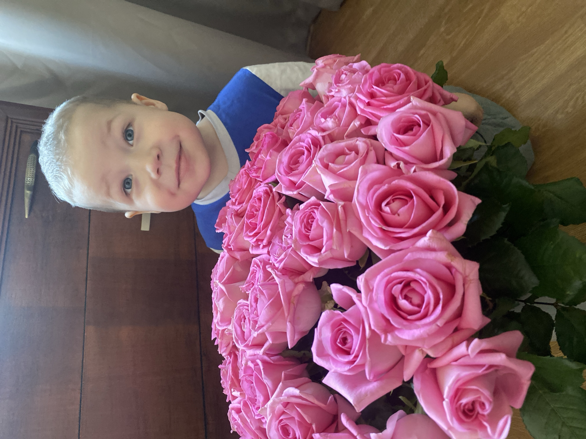 A young child with fair hair and blue eyes is positioned behind a large bouquet of pink roses. The child is wearing a blue and white striped shirt. To the left, a wooden door with a brass handle is visible. The background appears to be an indoor setting. The image provides no evidence of the location being in Sochi, Russia.