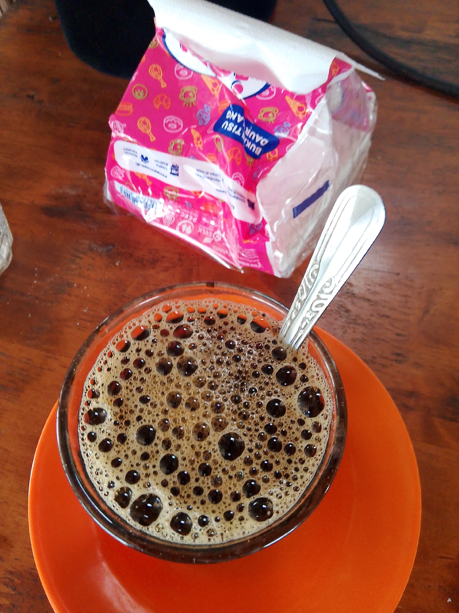 A close-up shot captures a glass of coffee with a foamy, brown surface and a silver spoon resting inside. The glass sits on an orange saucer, which is placed on a wooden table. In the background, a pink tissue packet with cartoon illustrations is partially visible. The scene appears to be set indoors, possibly at a cafe or a home in Meunasahmee, Indonesia.