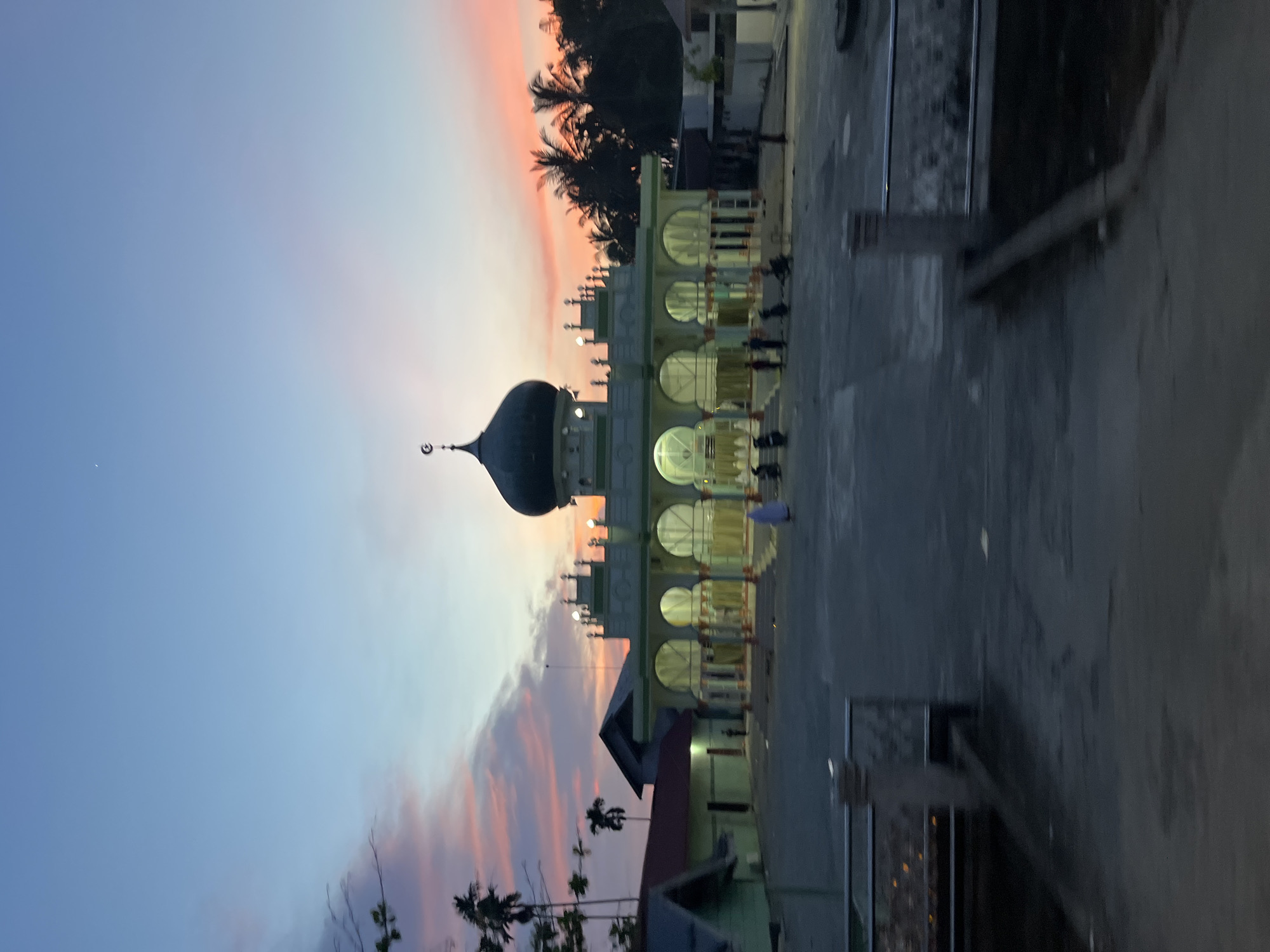 The image depicts a mosque in Matangpanyang, Indonesia, during sunset. The sky displays a gradient of blue, pink, and orange hues. The mosque features a prominent dark green dome topped with a crescent moon and star symbol. The building has a white facade with green accents and is supported by arches. Several people, some wearing light-colored clothing, are visible in front of the mosque, along with a few motorcycles. Palm trees and other vegetation surround the mosque, and the ground in front appears to be paved. The scene is illuminated by the twilight sky and some artificial lighting from the mosque.