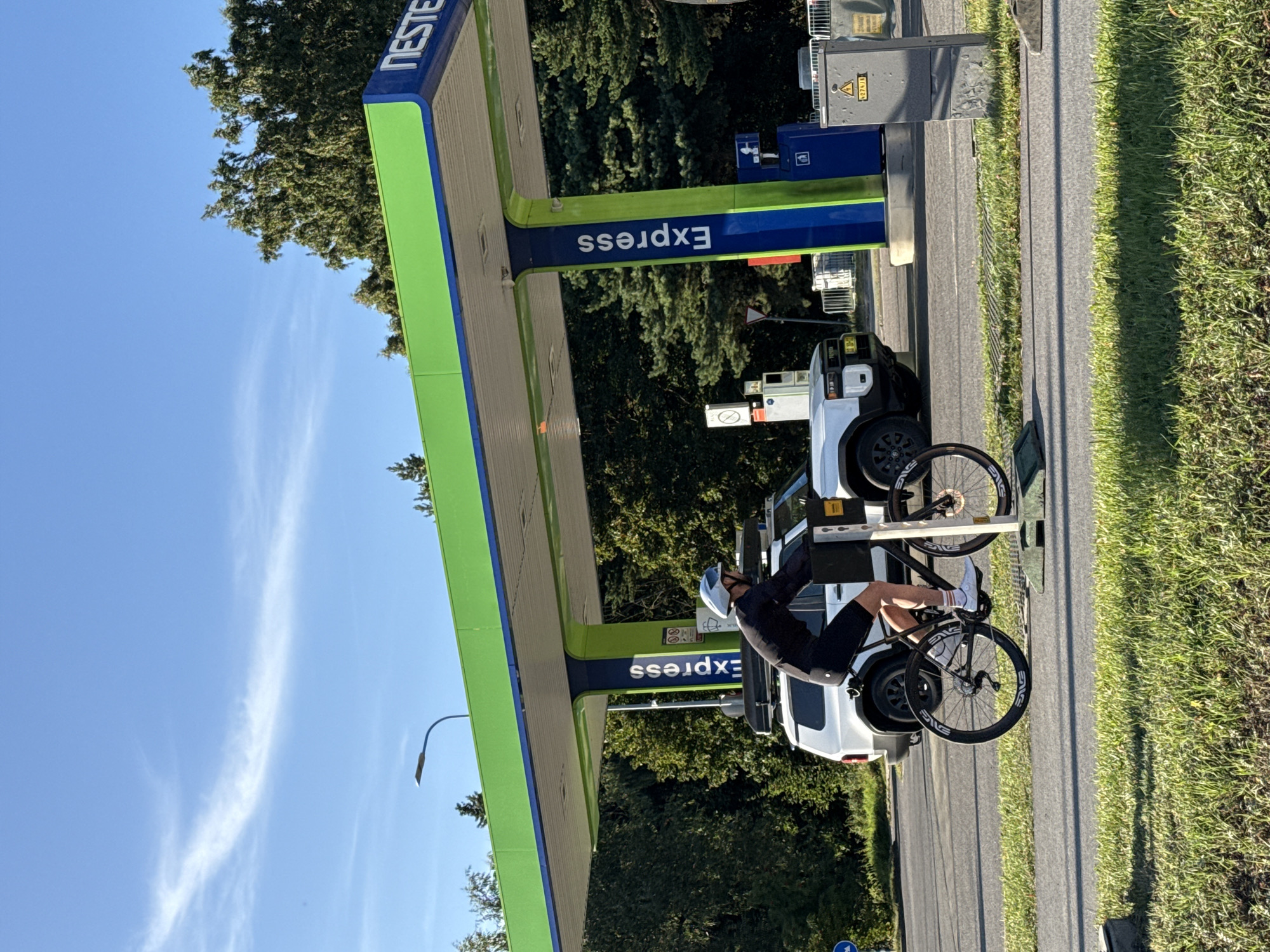 A man wearing a helmet, black long-sleeved shirt, and black shorts is cycling past a Neste Express gas station in Tallinn, Estonia. He is positioned on the right side of the frame, moving from left to right. A white SUV with a roof rack is parked under the gas station's green canopy. The gas station features blue pillars with white "Express" lettering. The sky is clear with a few wispy clouds. Trees line the background. The foreground shows a paved path and a grassy area.