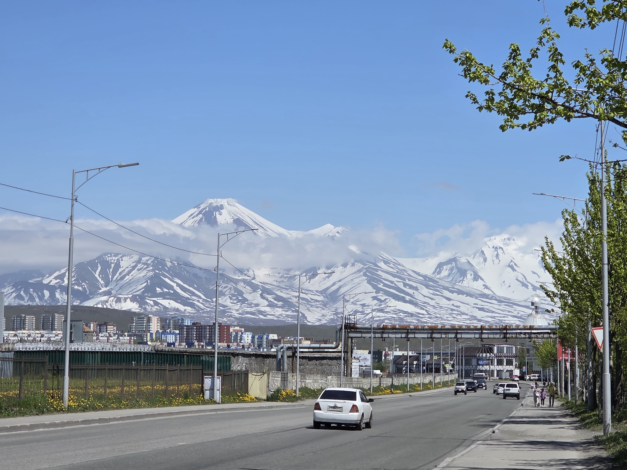 The image captures a bright, sunny day in a city with snow-capped mountains in the background. In the foreground, a paved road stretches into the distance, with a white car driving away from the camera. To the right of the road, a sidewalk is lined with trees and pedestrians walking. Further back, a row of buildings and industrial structures are visible, with a green fence running along the side of the road. The mountains, partially obscured by clouds, dominate the skyline. The overall scene suggests a developing urban area at the foot of a majestic natural landscape.