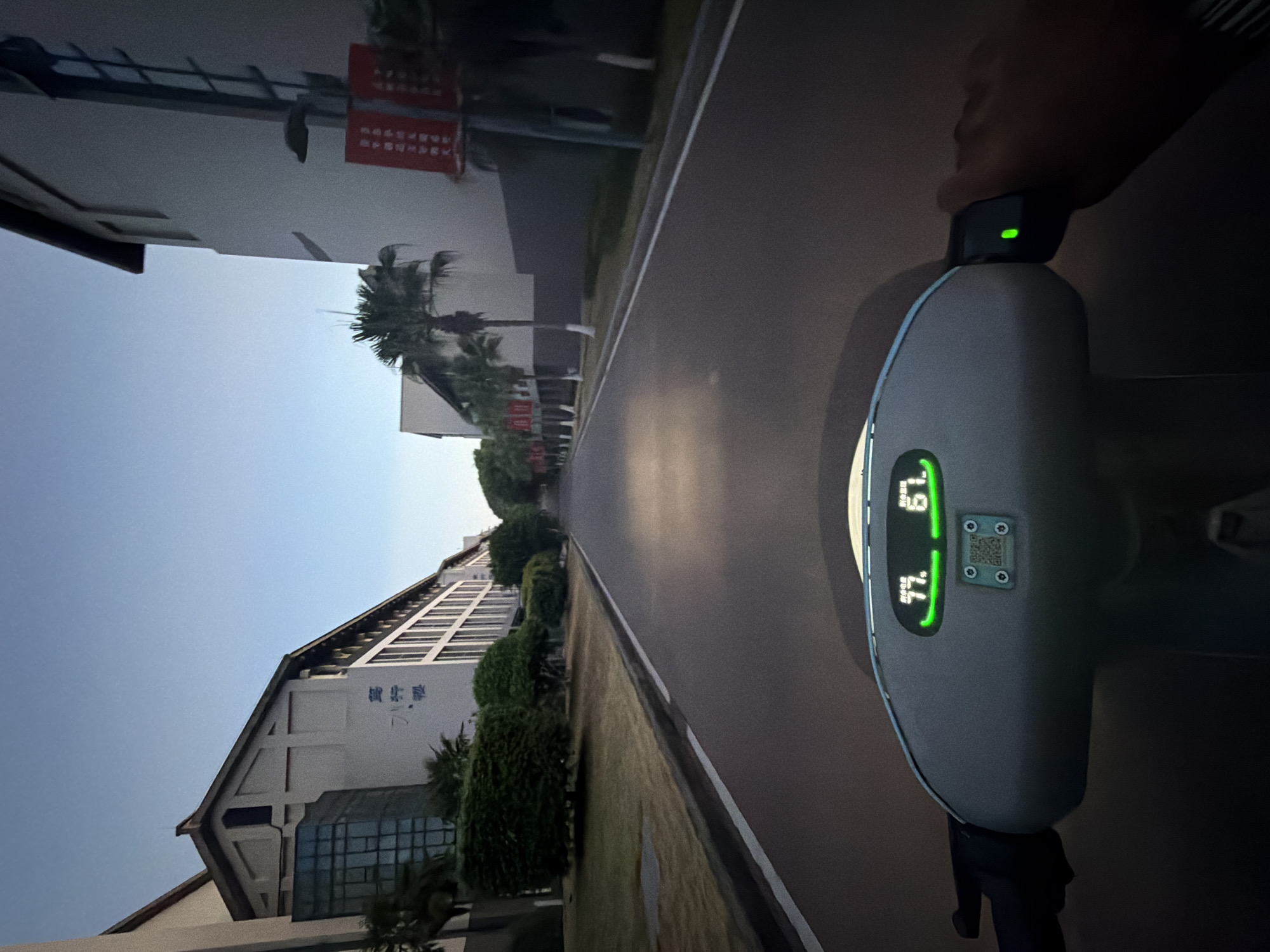 A first-person perspective shot shows the dashboard of an electric scooter in motion on a road. The scooter's dashboard displays two green digital readouts: "77." on the left and "61." on the right. A QR code is also visible on the dashboard. The road ahead is empty, bordered by manicured green bushes and trees on the left, and buildings with palm trees in front on the right. The sky is a clear, pale blue, indicating either early morning or late evening. The buildings appear to be part of a university campus or research facility, as indicated by Chinese characters on the facade of the building on the left, and red banners with text on the building on the right. The scooter is on a paved road within the city of Jiangjin, China.