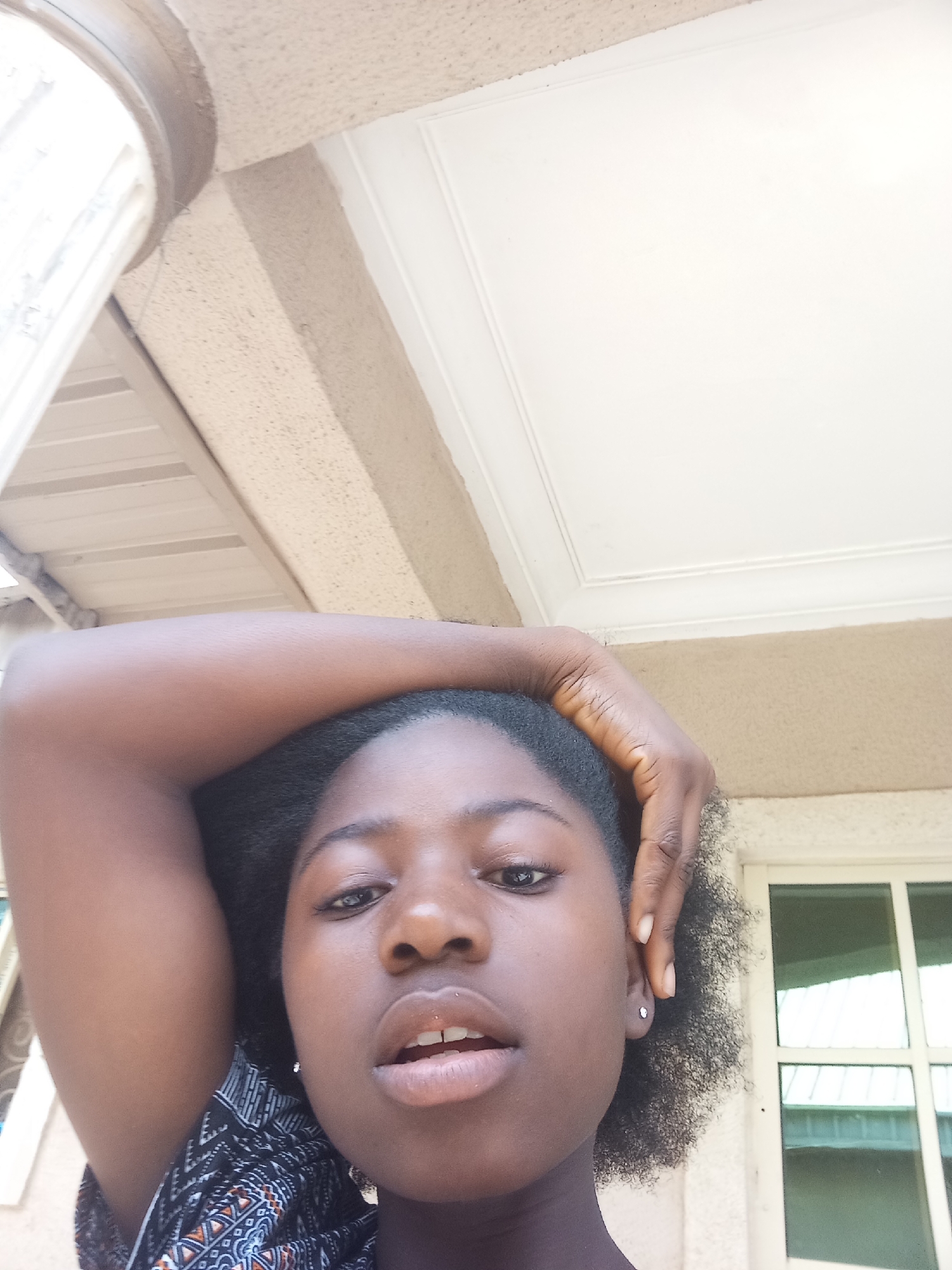 The image is a close-up, selfie-style photograph of a young Black woman. She is looking directly at the camera with a neutral expression. Her hair is black and curly, held on top of her head with one arm. She appears to be wearing a patterned top.
The background shows architectural elements, possibly a building or porch. There's a white ceiling with molding and a window with a grid pattern. The location is presumed to be Barikin Jirga Summa, Nigeria, but this is not explicitly verifiable from the image.