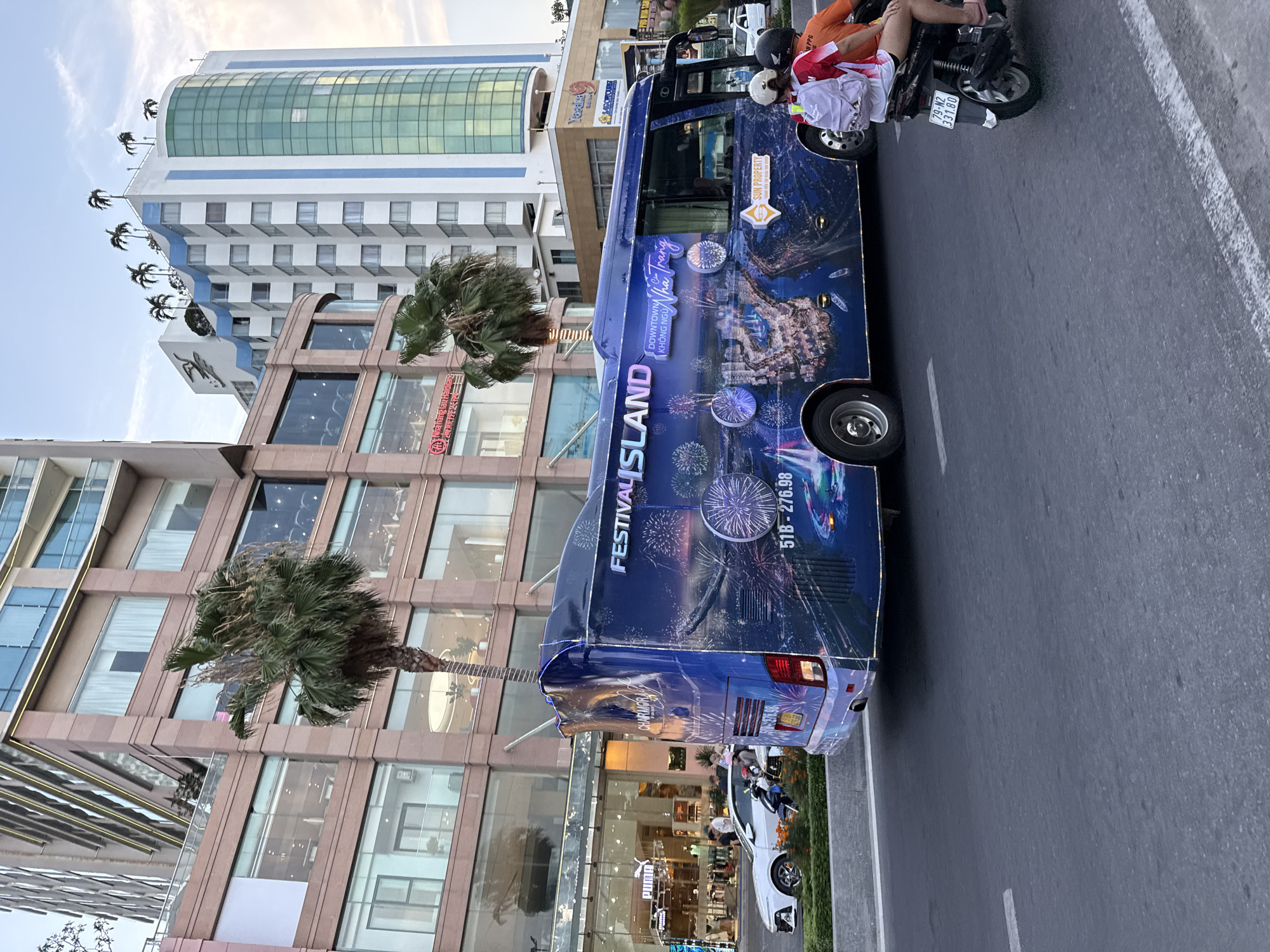 In Nha Trang Ward, Vietnam, a large blue bus decorated with images of fireworks and cityscapes drives down a paved road. The bus features the text "FESTIVAL ISLAND" and "DOWNTOWN KHÔNG NGỦ Nha Trang". On the side of the bus, a logo for "SUN PROPERTY" is visible. In the foreground, a motorcycle carrying two passengers, one with a white backpack and another wearing an orange shirt, travels alongside the bus. In the background, modern buildings with glass facades and palm trees line the street. A white car is parked on the side of the road near a building with a "Puma" store. The sky is overcast.
