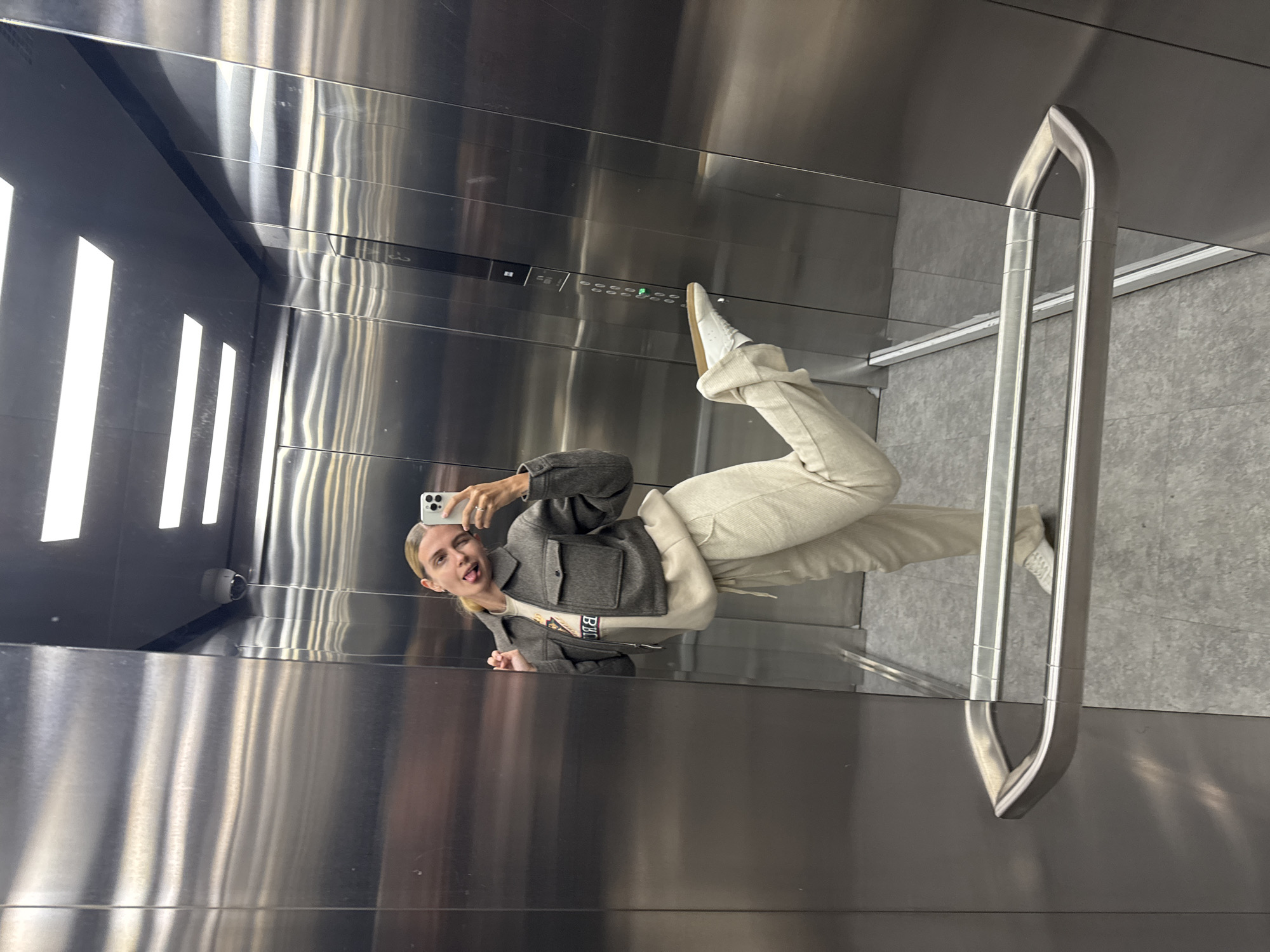 A person is reflected in the mirrored wall of a stainless steel elevator. The person is holding a smartphone and appears to be taking a selfie. The person is wearing a dark grey jacket over a light-colored sweatshirt, and light-colored pants and sneakers. The elevator has a metal handrail. The control panel with floor selection buttons is visible above the person's foot. The interior of the elevator is lit by vertical light panels. The location is an elevator in the city of Unknown, Unknown.