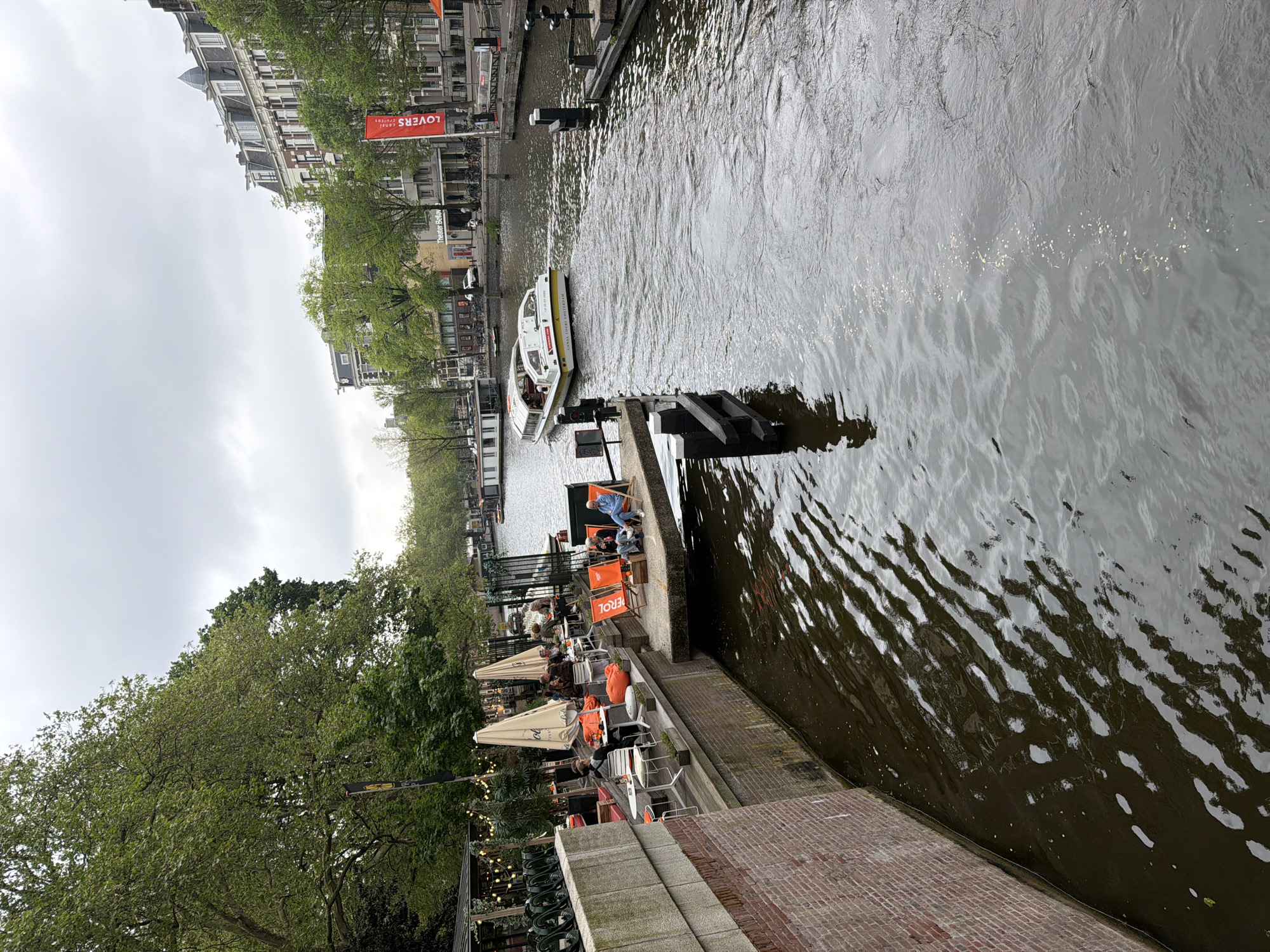 A canal in Amsterdam, Netherlands is viewed from a high angle on the edge of the canal. The water is dark and rippled. On the canal edge, there is outdoor seating for a restaurant with umbrellas, tables, and chairs. People are sitting and interacting. A small passenger boat is traveling down the canal. Along the opposite side of the canal, there are buildings and trees. The sky is overcast. A red sign that reads "LOVERS canal cruises" is visible along the opposite canal bank.