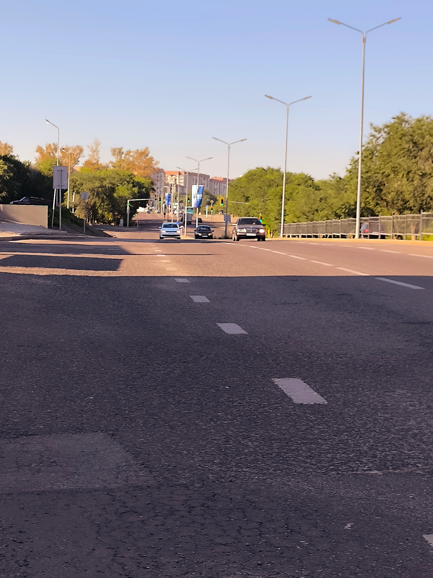 A wide, multi-lane paved road in Qarağandı city administration, Kazakhstan, is captured at a low angle. The road stretches into the distance, with white dashed lines marking the lanes and solid white rectangles indicating pedestrian crossings. The sky above is a clear, pale blue, with a few distant buildings visible in the background, suggesting an urban setting. Several streetlights, characterized by their tall, slender poles with V-shaped fixtures, line the sides of the road. Two cars, a white sedan and a dark-colored SUV, are driving in the same direction on the road. One car is in the lane closest to the viewer, while the other is further ahead. Green traffic lights are visible at an intersection in the distance, indicating that traffic is moving. The scene is bathed in warm, soft light, possibly from a sunrise or sunset, casting long shadows across the road. Lush green trees and some autumn-colored foliage line the sides of the road, with a metal fence bordering the right side. The overall impression is one of a peaceful urban environment during a calm period of the day.