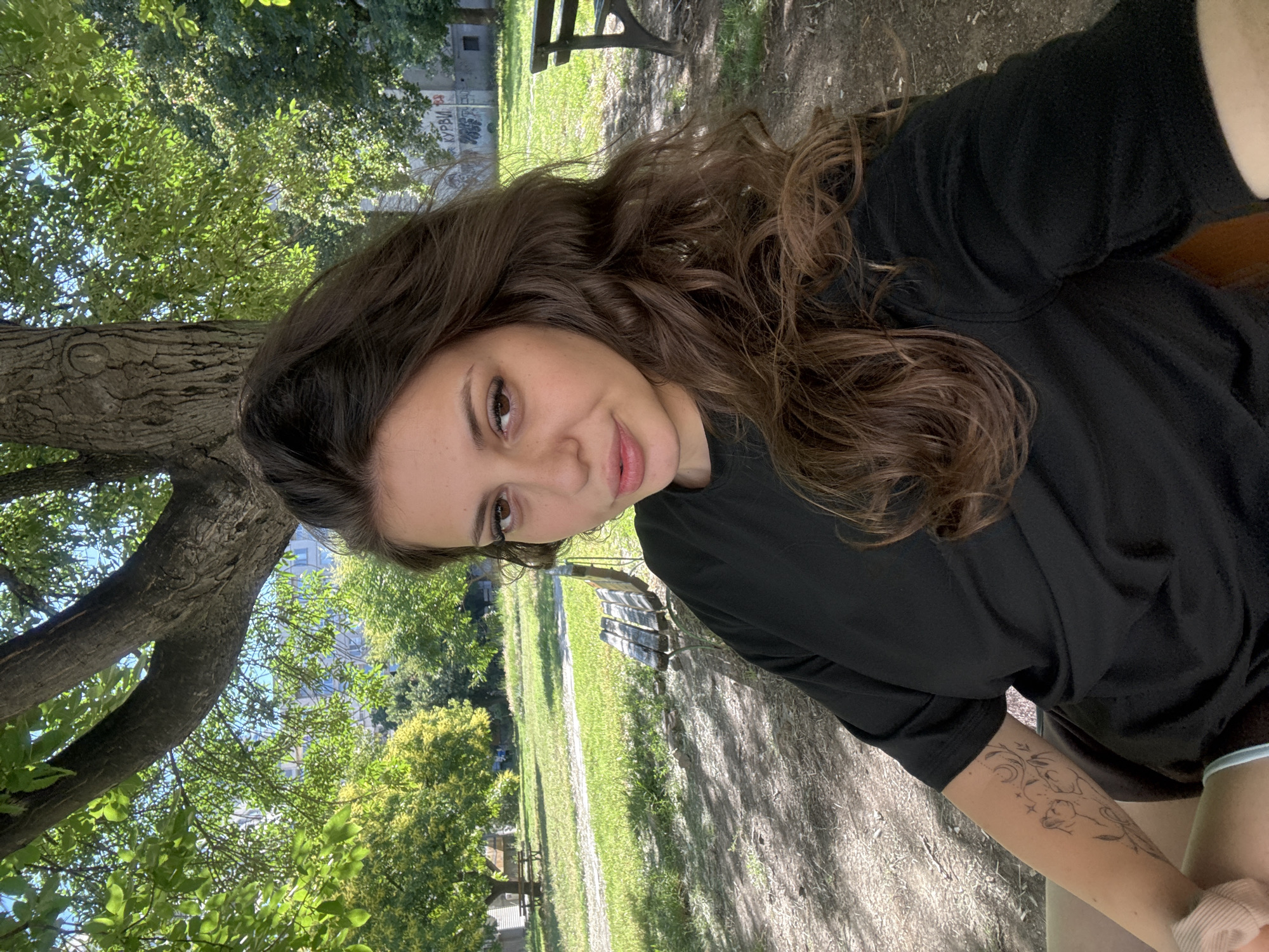 The image shows a young woman with long, brown, wavy hair, sitting in a park. She is wearing a black t-shirt and has a tattoo on her left arm. In the background, there are trees, green grass, and park benches. The scene suggests an outdoor setting, likely during daylight hours. The presence of graffiti on a building in the background is also noted.
The park setting and the presence of graffiti are consistent with urban environments. However, without further information or visual cues specific to Plovdiv, Bulgaria, such as distinct landmarks or architectural styles, it is not possible to definitively confirm the location as Plovdiv.