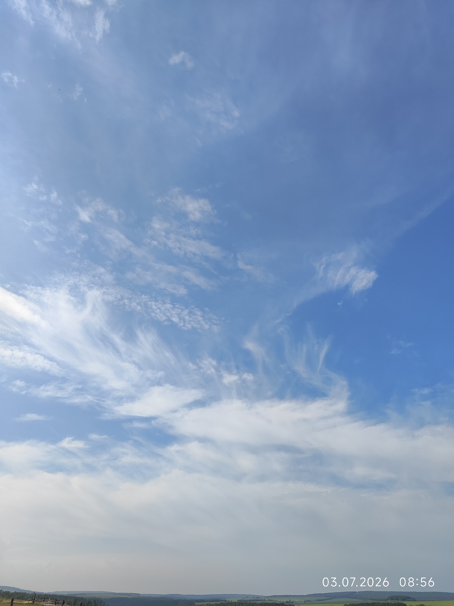 The image depicts a vast expanse of blue sky filled with wispy white clouds. In the lower portion of the frame, a hazy landscape is visible, characterized by rolling hills and scattered trees. A wooden fence runs along the very bottom left edge of the image. In the bottom right corner, white text displays the date "03.07.2026" and the time "08:56". The scene is a natural outdoor environment under a partly cloudy sky.