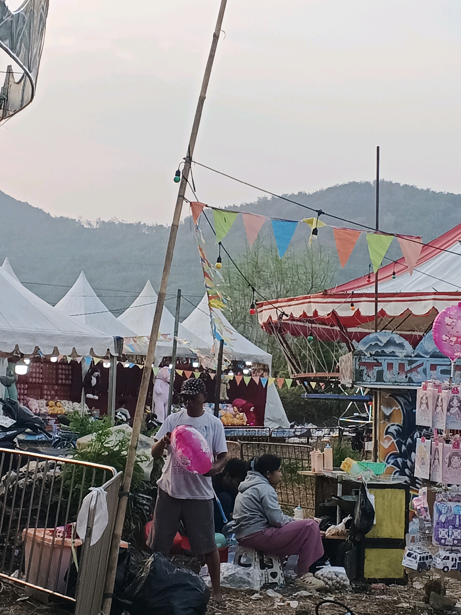 The image depicts an outdoor market scene, likely in Sukabumi, Indonesia, given the context. In the foreground, a man in a white t-shirt and patterned cap is holding a large, pink, heart-shaped balloon. He stands near a metal barrier and a trash bag. Behind him, to the left, several white tents with pointed roofs are set up, suggesting stalls or vendors. In the middle ground, a woman in a gray hooded sweatshirt and pink pants is sitting on a stool, looking to her right. She is near a small stand with bottles and other items. To the right, a red and white striped tent with decorative trim and flags is visible. Various goods, including what appear to be toys or merchandise, are displayed on stands and hanging from structures. A mountainous landscape forms the background under a hazy sky. The ground is covered in dirt and debris, indicative of an unpaved market area.