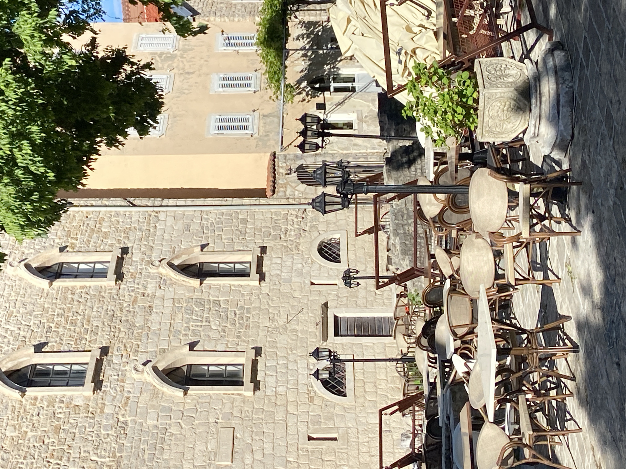 An outdoor scene in what appears to be a historic European town, characterized by stone buildings and a street lined with cafe furniture. The image shows a multi-story building constructed of light-colored, irregularly shaped stones, featuring several arched windows with stone frames. Below these, there are also some rectangular windows. Adjacent to this, another building with smooth, light-colored plaster walls is visible, distinguished by multiple windows fitted with white louvered shutters. A large green tree partially obscures the upper left portion of this building.
In the foreground and midground, numerous cafe tables and chairs are stacked, indicating that the establishment is either closed or preparing for operation. The chairs are made of wood and wicker, while the tables have light-colored circular tops. Several ornate, black streetlights with multiple lanterns are positioned along the street, casting shadows. A large, decorative stone planter with a green leafy plant is also visible among the stacked furniture. Overhead, a large, cream-colored fabric awning is partially unfurled.
The ground is paved with cobblestones or pavers. Bright sunlight illuminates the scene, casting distinct shadows from the buildings, streetlights, and stacked furniture, suggesting it is daytime, likely morning or late afternoon. No people are visible, contributing to a quiet and serene atmosphere. There is no visible text in the image.