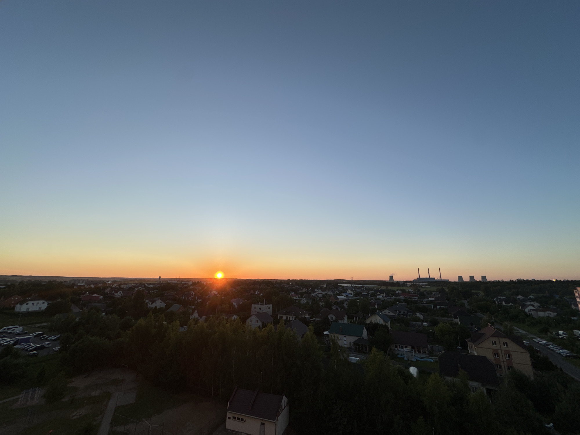 This image captures a wide-angle, aerial view of a suburban landscape in the Shchomyslicki Sel'ski Savet, Belarus, during sunset. The sun, a small, bright orange orb, is visible low on the horizon, casting a warm glow across the scene. The sky transitions from a pale blue at the top to a soft orange and yellow gradient near the horizon.
The foreground is dominated by dense, dark green trees, obscuring much of the ground below. A small, light-colored building with a dark roof is partially visible among the trees in the bottom center of the frame. To the left, a parking lot filled with numerous cars is visible, suggesting residential or commercial activity.
Beyond the trees, a sprawling residential area unfolds, characterized by numerous houses with varying roof colors and architectural styles. The houses are interspersed with more trees and patches of green. In the distance, a line of industrial structures with tall chimneys is silhouetted against the horizon, hinting at a power plant or industrial complex. A few scattered buildings and structures are also visible further out. The overall impression is one of a developing suburban area with a mix of residential housing and distant industrial elements. There are no people or animals visibly present in the image. The lighting suggests late evening, with the sun nearing the end of its diurnal path.