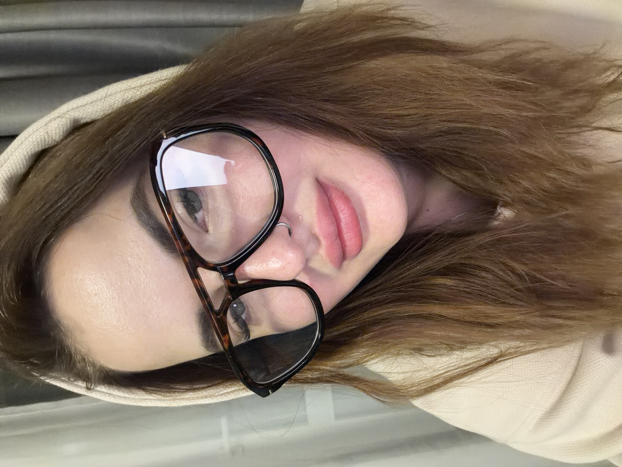 A light-skinned woman with brown hair and wearing eyeglasses, a nose ring, and a beige hoodie, is seen in a selfie, turned sideways. The background is a greyish-beige fabric. There is no indication of the location being in Krasnodar, Russia.