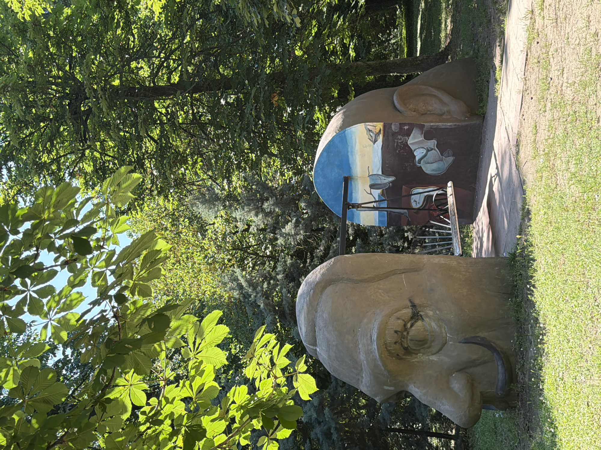 The image depicts a surrealist sculpture inspired by Salvador Dali's "The Persistence of Memory" located in Pazardzhik, Bulgaria. The sculpture consists of two large, stylized human heads. The head on the left is a profile view, featuring a prominent nose, eye, and a mustache resembling Dali's signature look. The head on the right is also a profile, but its inner surface is painted with a scene from "The Persistence of Memory," complete with melting clocks and a landscape. A bench is positioned between the two heads, suggesting it's part of the artistic installation. The sculpture is set in a park-like environment with lush green grass, trees, and sunlight filtering through the leaves. The overall scene is artistic and whimsical, designed to evoke the surrealist style of Salvador Dali.