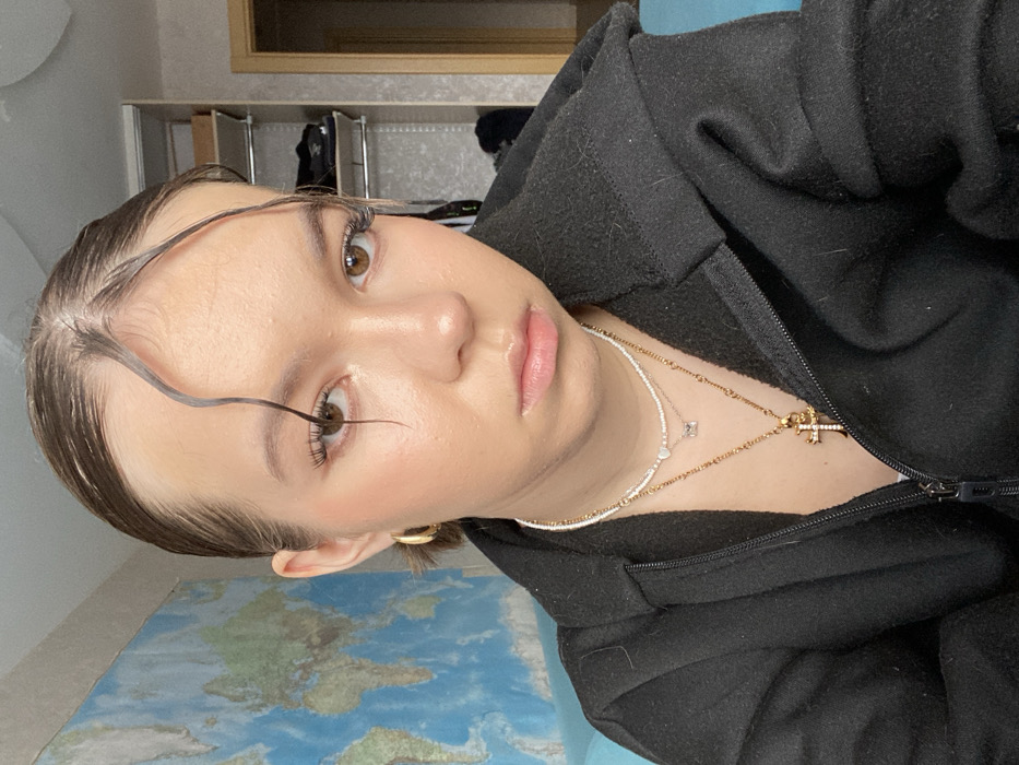 A female individual is depicted in a close-up, head-and-torso shot, facing and looking directly into the camera. The setting is an indoor residential room in Chaikovsky, Russia. She has a light complexion, brown eyes, and dark brown hair styled with a central part and two distinct wet-look wavy strands framing her forehead. She exhibits light cheek blush and mascaraed eyelashes. Jewelry includes small gold hoop earrings, a white beaded choker with a small heart charm, a thin gold-colored chain with a rectangular clear stone pendant, and a thicker gold-colored chain with an ornate gold-colored cross pendant. She is wearing a black zippered fleece or hooded jacket, partially unzipped at the front. The background on the left features a large, predominantly blue world map poster on a wall. On the right, a light-colored shelving unit containing dark items, possibly clothing, is visible, along with a light-colored door frame. A blue fabric surface, likely a sofa or chair, is visible at the lower left and right edges of the frame.