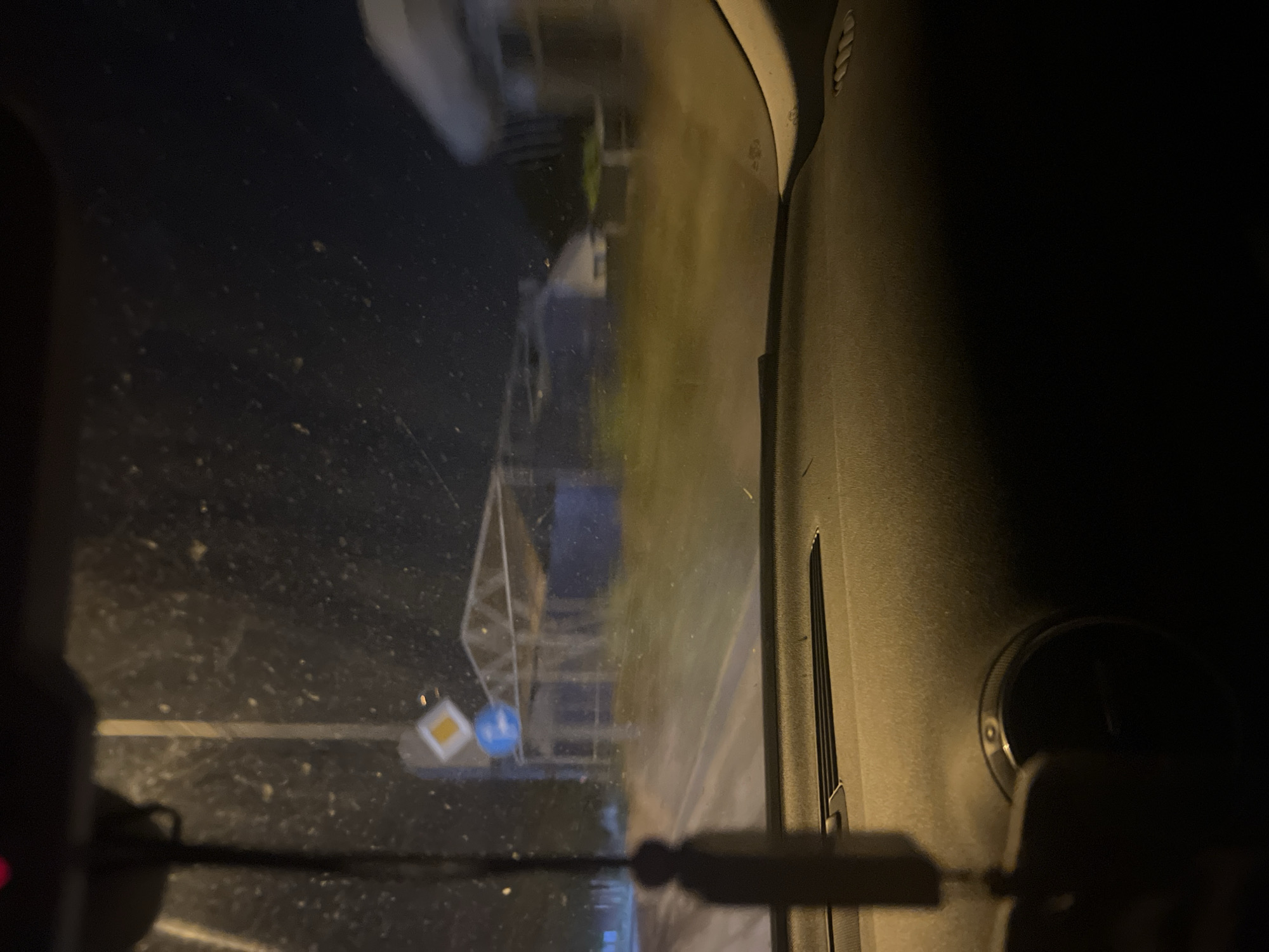 The image depicts a view from inside a vehicle at night, looking out through the windshield. The windshield is dirty, with visible specks and smudges. In the foreground, a dark dashboard is visible, with a circular vent on the right side. Hanging from the rearview mirror is a dark tassel-like ornament.
Outside the vehicle, a street is visible, with a lamp post and traffic signs. One sign is a diamond shape with a yellow center, and below it is a blue circular sign. Further down the street, there appears to be a covered structure, possibly a bus stop or a small shelter, with a metal frame and a translucent roof. Beyond this structure, indistinct buildings or houses are visible, illuminated by streetlights. The environment is dark, suggesting it is late at night, and the ground is wet, possibly from rain or recent watering. The overall impression is a blurry, nighttime street scene, likely in a residential or semi-urban area, given the presence of houses and the covered structure. The location is identified as Krivina, Bulgaria.