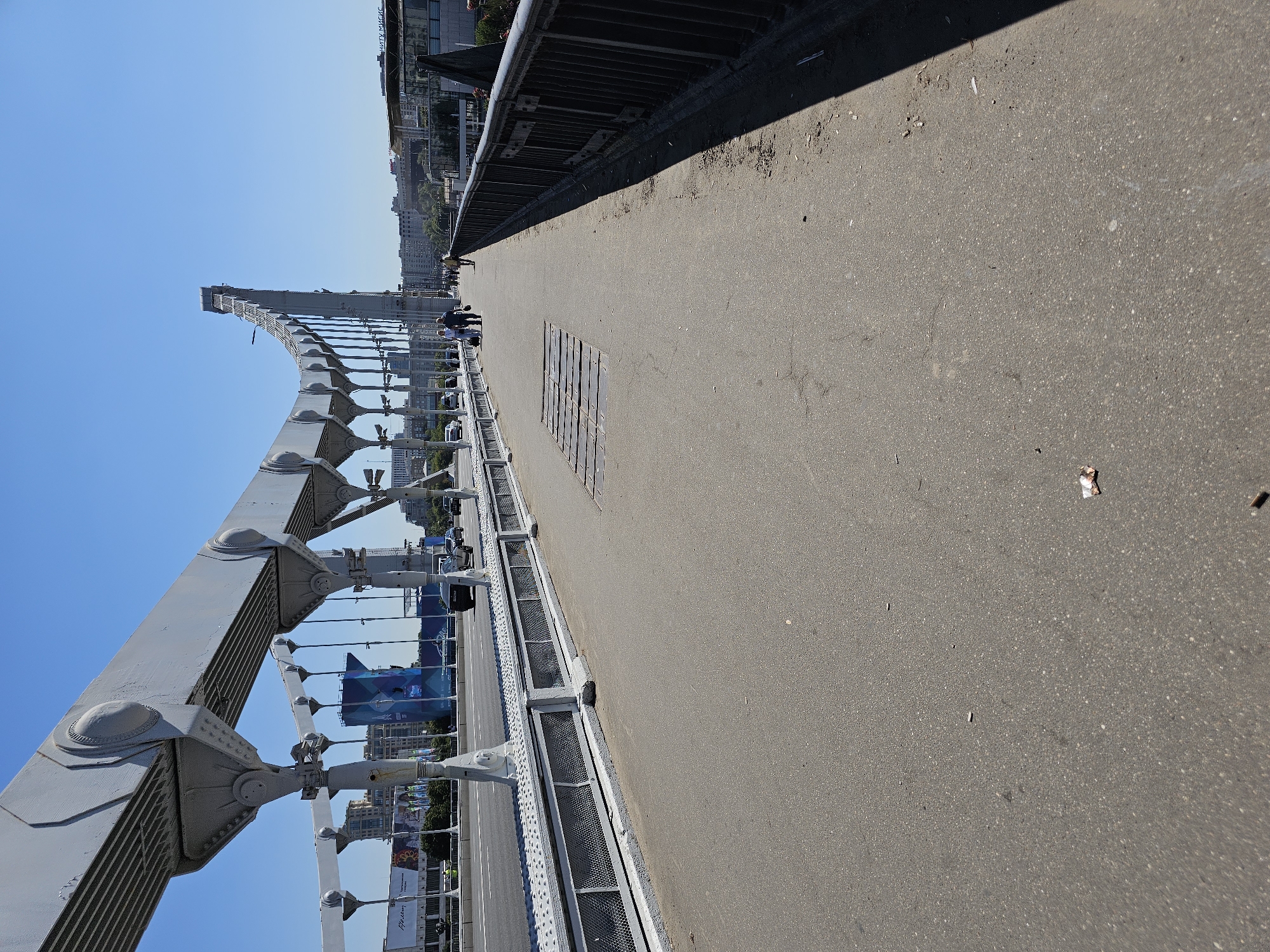 The image shows a view from a pedestrian walkway on a suspension bridge on a clear, sunny day. The walkway is paved with asphalt and runs alongside a roadway with cars in motion. On the left, a large gray suspension structure of the bridge dominates the frame with its cables and supporting beams. In the distance, various buildings and a large blue advertisement banner are visible. Several pedestrians are walking on the bridge, some with luggage. A black car is seen on the road. The railing of the pedestrian walkway is dark and metal. The ground of the walkway has some scattered debris, including cigarette butts and wrappers. The sky is clear blue.