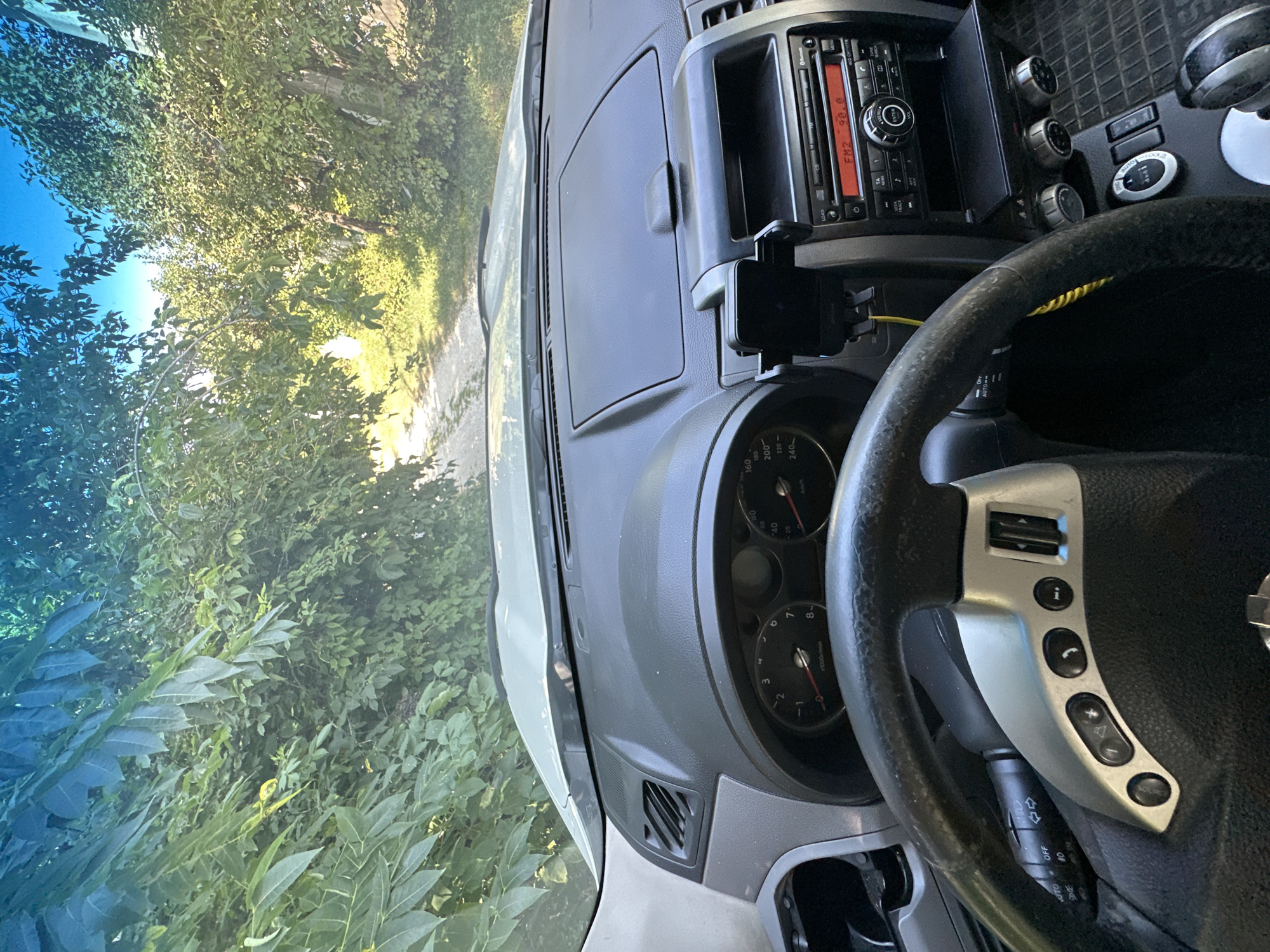 The image shows the interior of a car parked on a dirt road surrounded by lush green trees and foliage. Sunlight filters through the leaves, creating dappled light. The dashboard is visible, including the steering wheel, instrument cluster, and car stereo. The radio display shows "FM2 90.0". The car appears to be parked in a rural area. The location is specified as Makiv, Ukraine.