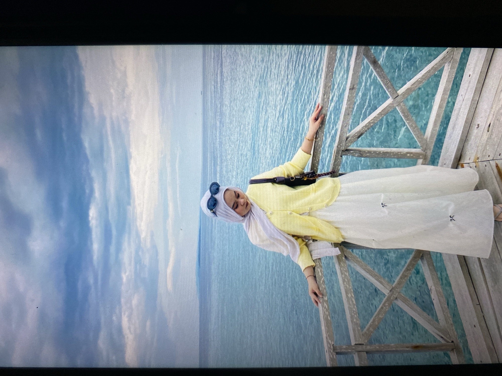 A woman wearing a light yellow cardigan, a white skirt, and a light blue hijab stands on a wooden pier overlooking the ocean. She has sunglasses perched on her head and a dark shoulder bag. The ocean is a vibrant turquoise, and the sky is filled with dramatic clouds. The pier is made of weathered wood with crisscrossing railings. The background shows a hazy horizon with a distant landmass. The location is in Ta', Indonesia.