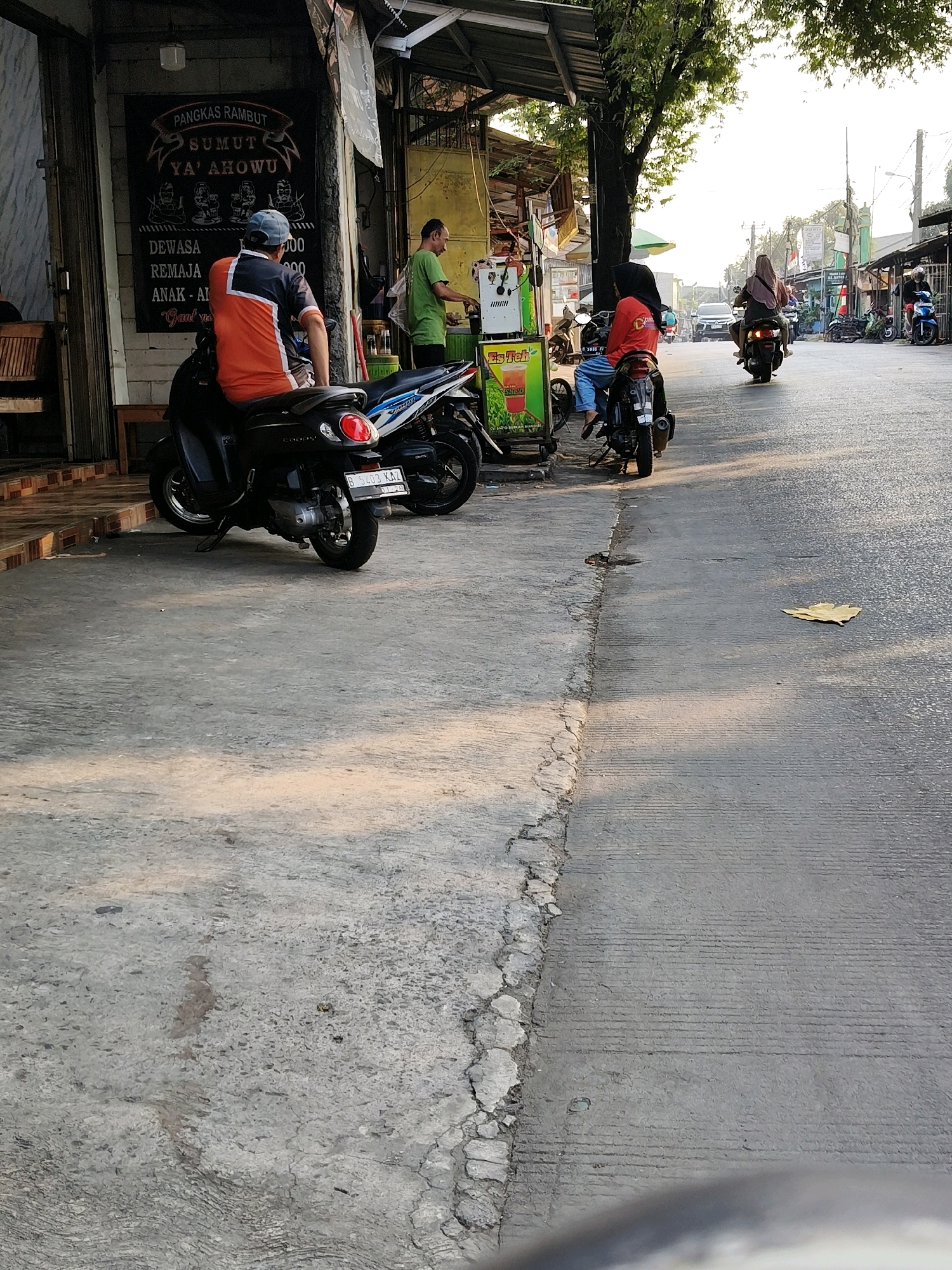 A street scene in Bekasi, Indonesia, depicts a commercial area with several motorcycles and people. In the foreground, a man wearing a blue cap and an orange and black shirt sits on a black motorcycle. Next to him, another black motorcycle is parked. To the left, a building with a sign for "PANGKAS RAMBUT SUMUT YA' AHOWU" (a barbershop) is visible, with wooden chairs and tables outside. Further down the street, a woman in a red hijab and an orange shirt is on a motorcycle, and behind her, another woman in a brown hijab rides a motorcycle. Several other motorcycles are parked along the street, and a food cart selling "Es Teh" (iced tea) is also present. The street is paved and shows signs of wear. Trees line the right side of the street, and the sky is bright with late afternoon sun.