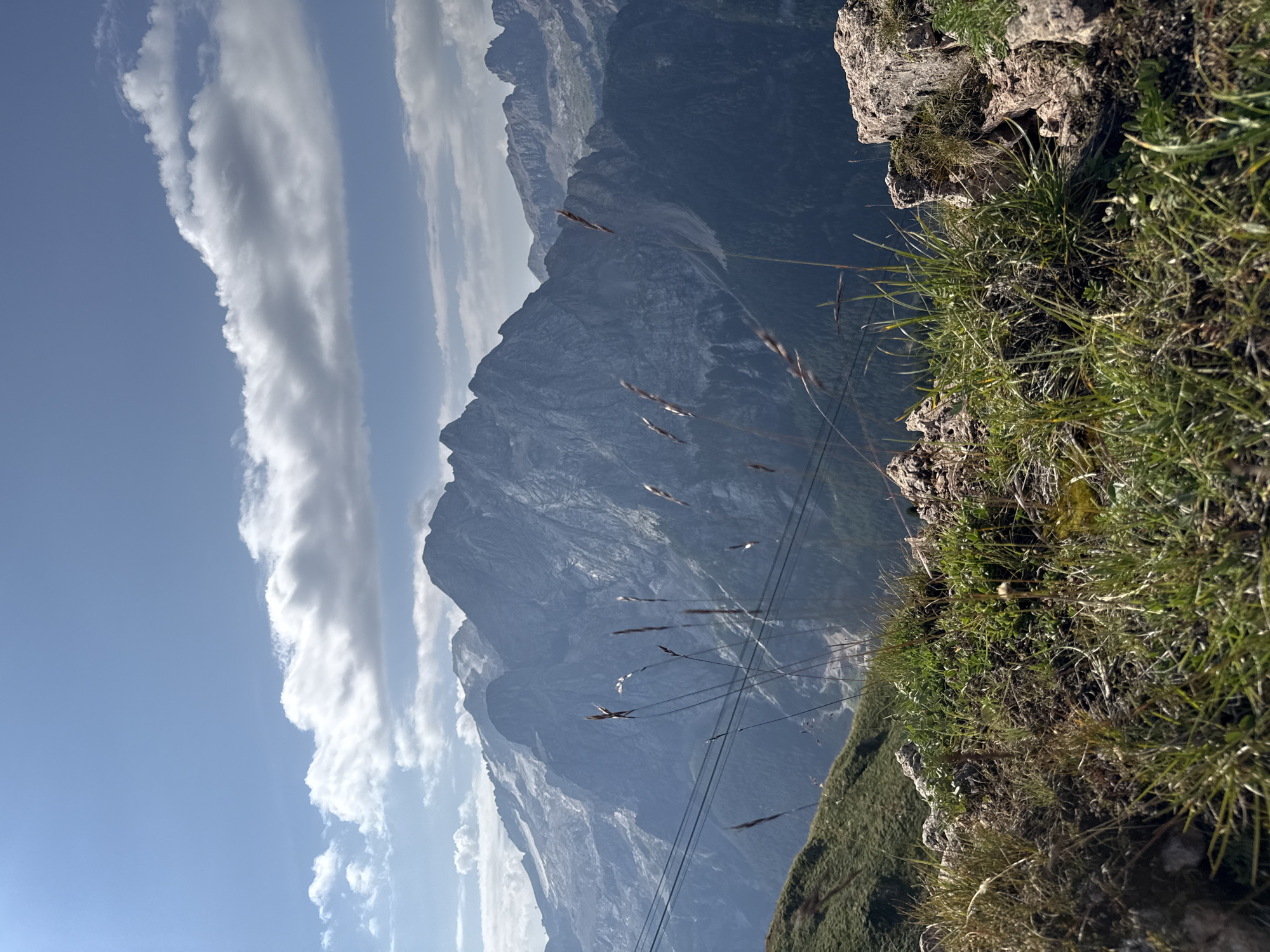 This is a scenic outdoor photograph taken from a high vantage point in the Dolomite mountains, likely near Canazei, Italy, given the characteristic peaks. The foreground is dominated by a rocky, grassy slope with dry grasses and small green plants. Several thin, dry stalks of grass are prominently featured in the center of the frame, some blurred due to their proximity to the camera.
In the midground, cable car wires stretch horizontally across the scene, supported by unseen pylons, suggesting a tourist or transport infrastructure. The wires cast subtle shadows on the grassy hillside. Beyond the wires, a vast mountain range unfolds. The mountains are rugged and imposing, with steep slopes and rocky faces. Some of the higher peaks appear to be capped with snow or glaciers, their surfaces reflecting the sunlight. The distant peaks are shrouded in a bluish haze, typical of atmospheric perspective.
The sky above is a clear, bright blue, dotted with scattered white clouds. Some clouds are wispy and thin, while others are larger and more voluminous, casting soft shadows on the mountains. The overall impression is one of natural grandeur and the vastness of the alpine landscape under a sunny sky. There are no people visible in the photograph.
