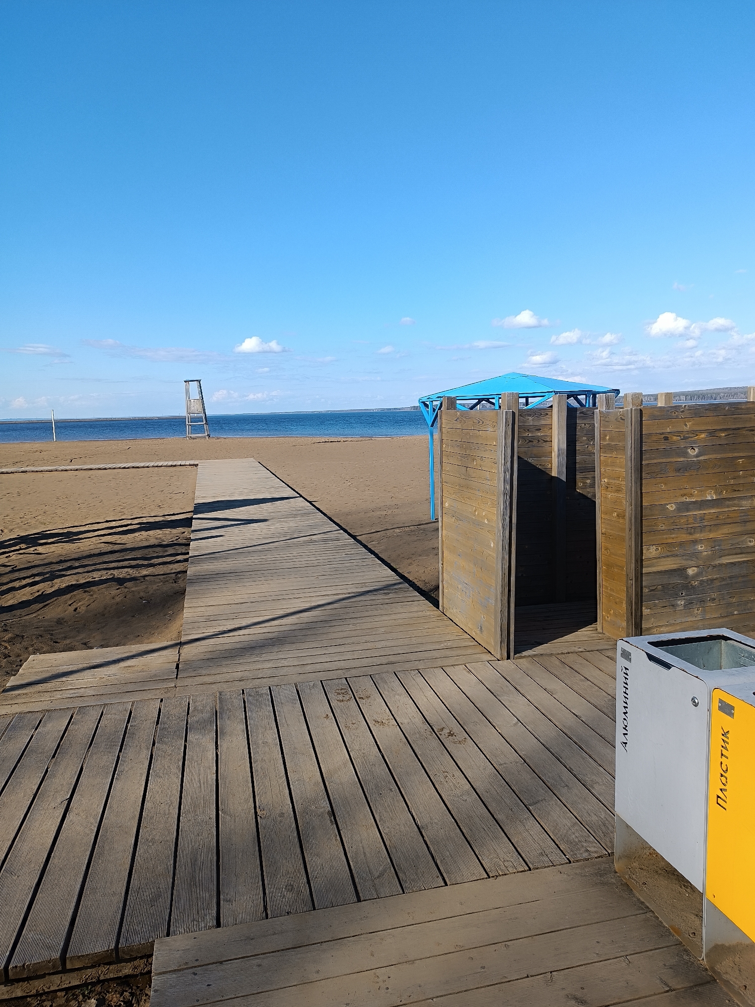 This image captures a tranquil beach scene, likely located in Chaikovsky, Russia, under a clear, bright blue sky dotted with scattered white clouds. The setting appears to be a prepared recreational area, devoid of people, suggesting a quiet moment.
In the foreground, a network of wooden boardwalks extends across the sandy beach. A primary boardwalk leads towards the distant body of water, with a branching path to the left. These wooden paths feature textured surfaces and show signs of weathering. Long shadows are cast from the boardwalk structures, indicating either morning or late afternoon sunlight.
To the right, two wooden changing cabins, constructed from dark brown planks, stand adjacent to the boardwalk. Behind them, a blue metal-framed canopy or pavilion provides shade. A modern recycling bin is partially visible on the far right, featuring silver and yellow sections. The visible Cyrillic text on the bin reads "АЛЮМИНИЙ" (ALUMINIUM) on the silver part and "ПЛАСТИК" (PLASTIC) on the yellow section.
In the midground, near the water's edge, a tall wooden lifeguard stand is visible. An expansive body of water, likely a lake or sea, stretches across the horizon. The absence of people contributes to a peaceful and inviting atmosphere, depicting a well-maintained beach on a pleasant, sunny day.