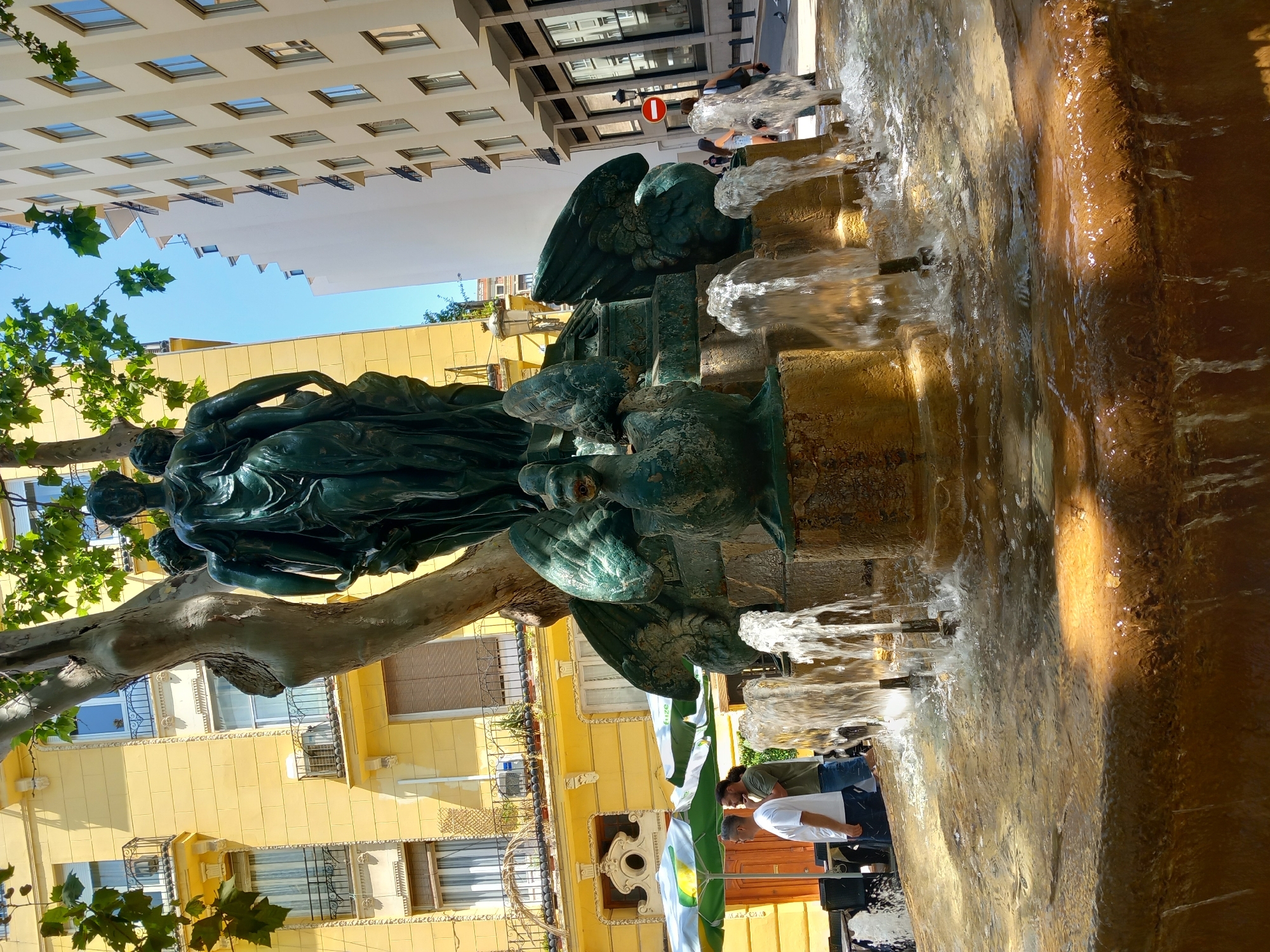 A bronze sculpture of figures and waterfowl sits atop a multi-tiered fountain with water spraying upwards and flowing over the sides. The sculpture is dark green with verdigris patina. The fountain base is made of weathered stone and concrete. In the background, a yellow building with balconies and windows is visible, along with a tall, light-colored building. Two men stand under umbrellas to the left of the fountain, one wearing a white t-shirt and the other a green t-shirt. A street with a red and white circular sign is visible to the right, with a few people walking in the distance. The scene appears to be a public plaza in the city of València, Spain, possibly in the Ciutat Vella district, given the architectural style and the presence of a historic fountain.