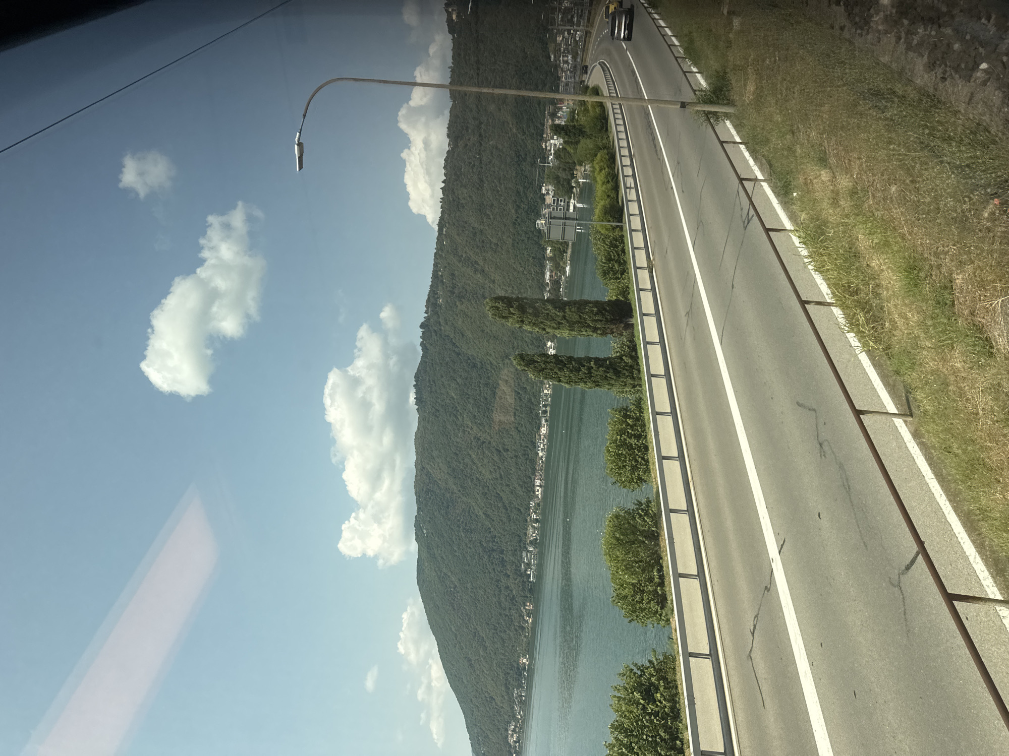 The image shows a scenic view of a road alongside a lake, likely taken from a moving vehicle. The road curves to the right, with two lanes separated by a white line. A metal guardrail runs along the side of the road, separating it from a grassy embankment. Across the lake, a densely forested hill rises steeply, dotted with buildings that form a settlement. The lake itself is a deep blue-green, and the sky above is a clear blue with scattered white clouds. A street lamp stands on the right side of the road, and a few cars are visible on the road. In the foreground, the grass appears dry and somewhat overgrown. The reflection of something rectangular and pale, possibly from the interior of the vehicle, is visible in the upper left portion of the image, suggesting it was taken through a window. The overall impression is one of a picturesque landscape along a transportation route, likely in a town situated on the shores of a lake.