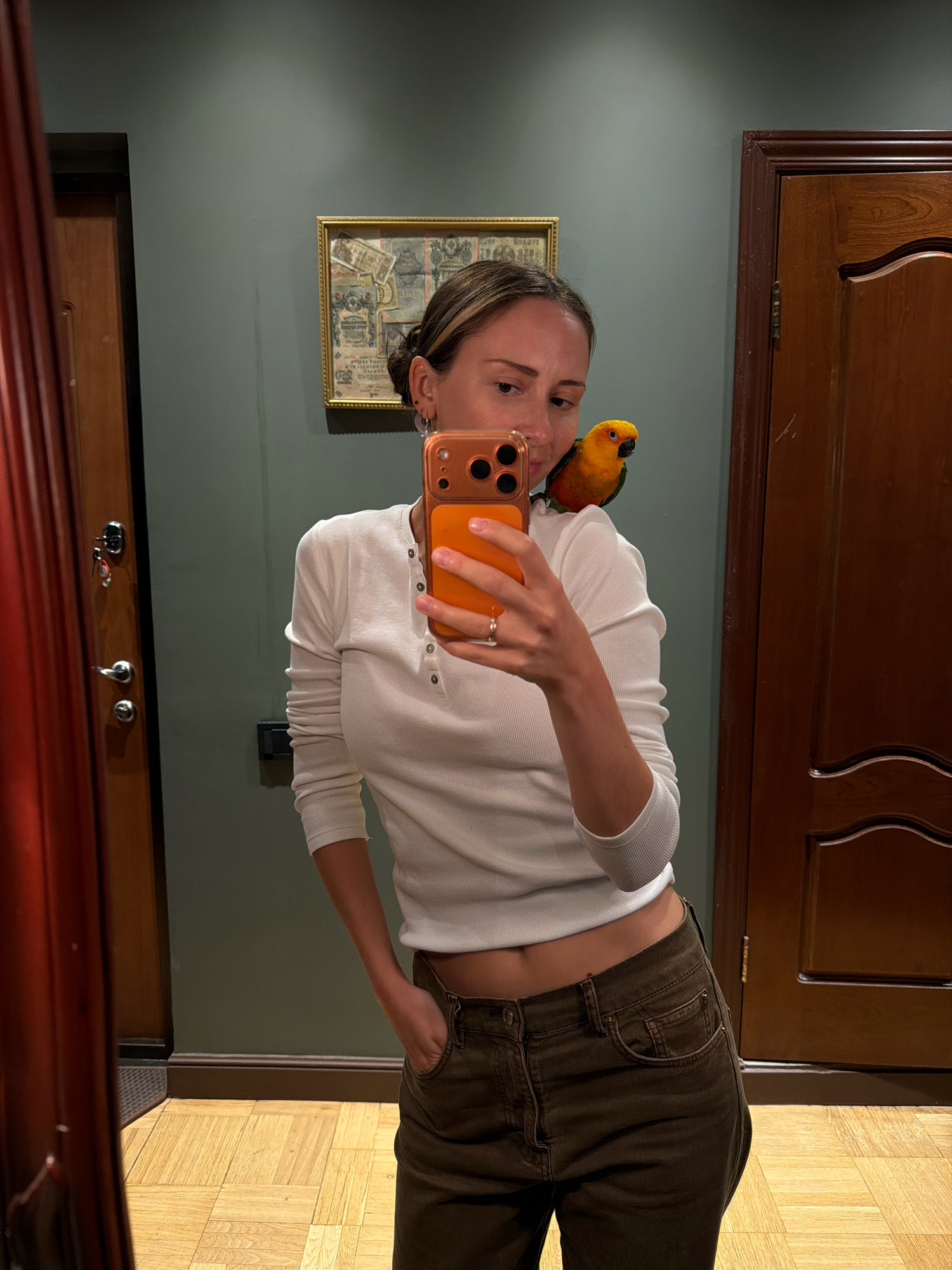 In a mirror selfie taken indoors, a woman stands with a small, vibrantly colored parrot perched on her right shoulder. The woman is wearing a white, long-sleeved ribbed shirt and dark brown pants, with one hand casually resting in her pocket. She is holding an orange smartphone in her left hand, angled to capture the reflection. The parrot, predominantly yellow with orange and green accents, looks towards the woman. The background features a dark green wall with a framed picture, a wooden door, and a reflection of a reddish curtain. The flooring is a light-colored wood laid in a parquet pattern.