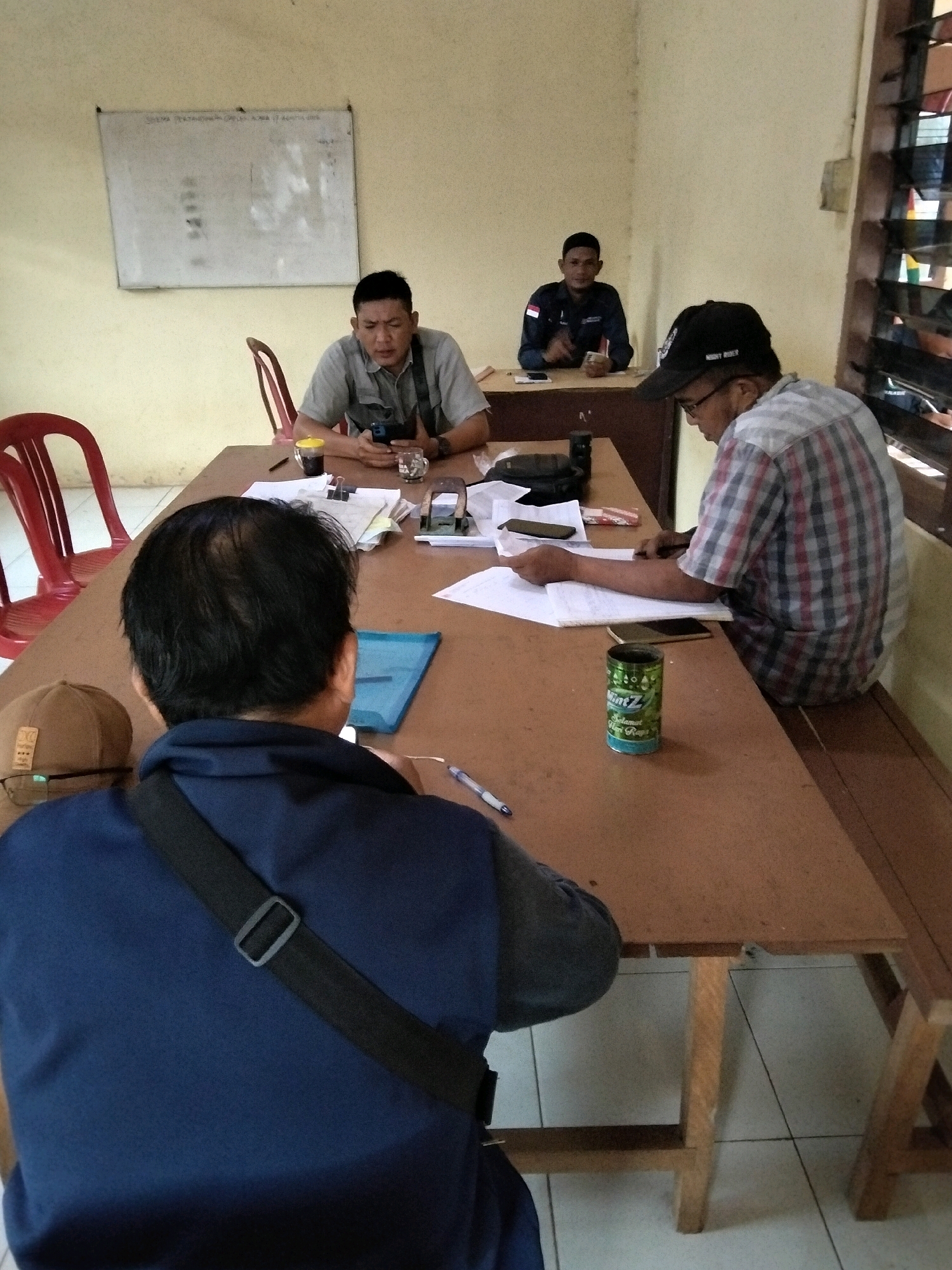 The image depicts an indoor scene, likely an office or meeting room, in Cinta Manis Lama, Indonesia. Four individuals are present, seated around a wooden table. In the foreground, a man with dark hair is seen from behind, wearing a navy blue shirt and a black shoulder bag. To his left, a man in a grey collared shirt is seated, looking at his phone. He has a yellow-lidded cup and a small glass on the table in front of him. Further back and to the right, a man wearing a checkered shirt and a black baseball cap is looking down at papers on the table. Behind the man in the grey shirt, another man in a dark blue collared shirt with an Indonesian flag patch is seated at a separate table, facing the camera and holding a document. The room has light-colored walls and a wooden table. A whiteboard with some writing is visible on the wall behind the man in the grey shirt. A red chair is positioned near the table. A can of "Mint Z" is on the table in front of the man in the checkered shirt. The floor appears to be tiled.