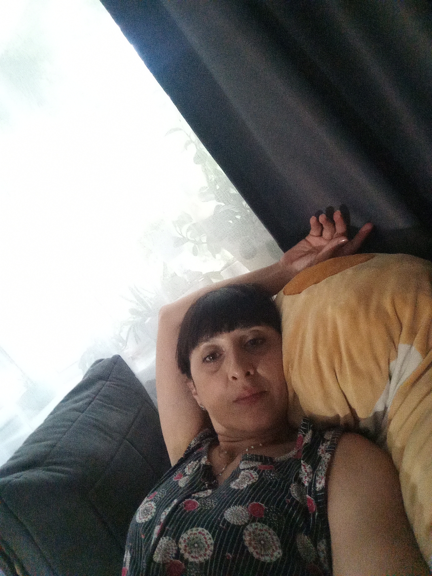 The image is a selfie taken by a woman with short, dark hair, wearing a dark dress with a floral pattern. She is lying down, with her right arm raised and resting behind her head. Her face is turned towards the camera, and she appears to be looking at it. She is positioned on a bed or sofa, with pillows visible behind and to her left. The background consists of a window with sheer curtains, allowing natural light to enter, and a dark curtain on the right side of the frame. The overall lighting suggests it is daytime. The scene appears to be indoors, likely a bedroom or living room. There is no indication of the specific location within Belogorsk, Russia.