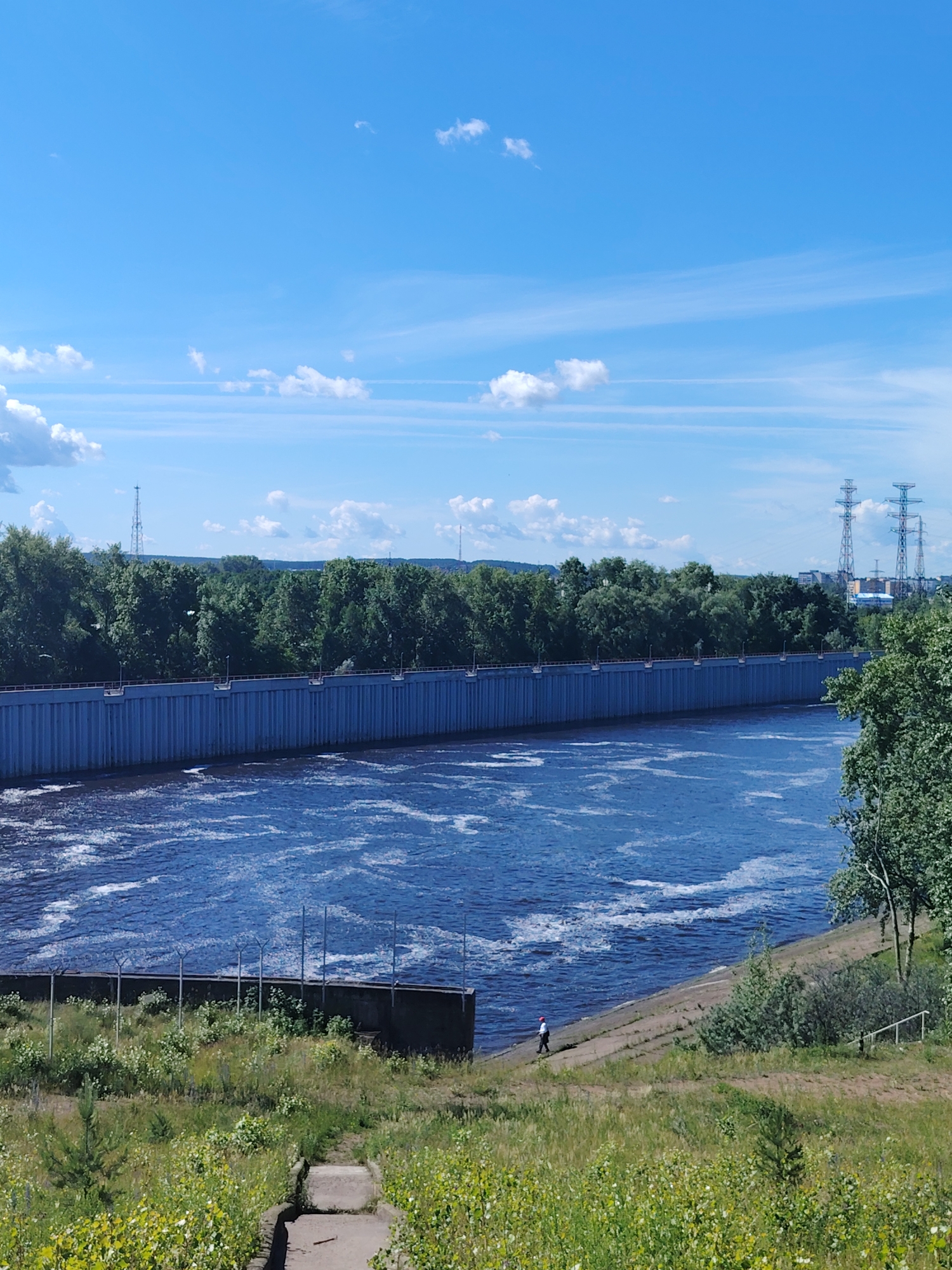 The image depicts a wide, turbulent body of blue water, likely a river, with white foam churning on its surface. A tall, blue concrete dam runs horizontally across the mid-section of the frame, separating the water from a densely wooded area beyond. The dam is reinforced with vertical structures and topped with a railing. On the bank of the river, closer to the viewer, is a grassy slope dotted with various green shrubs and some yellow wildflowers. A concrete path or stairway descends the slope. A lone individual, wearing a white shirt and red cap, is walking along the sloped embankment near the water's edge. In the background, beyond the trees, are high-voltage electricity pylons and distant buildings, suggesting an urban or industrial landscape. The sky is bright blue with scattered white clouds. This scene is consistent with the Kama River and the Chaikovsky Hydroelectric Power Station in Chaikovsky, Russia, a city on the Kama River.