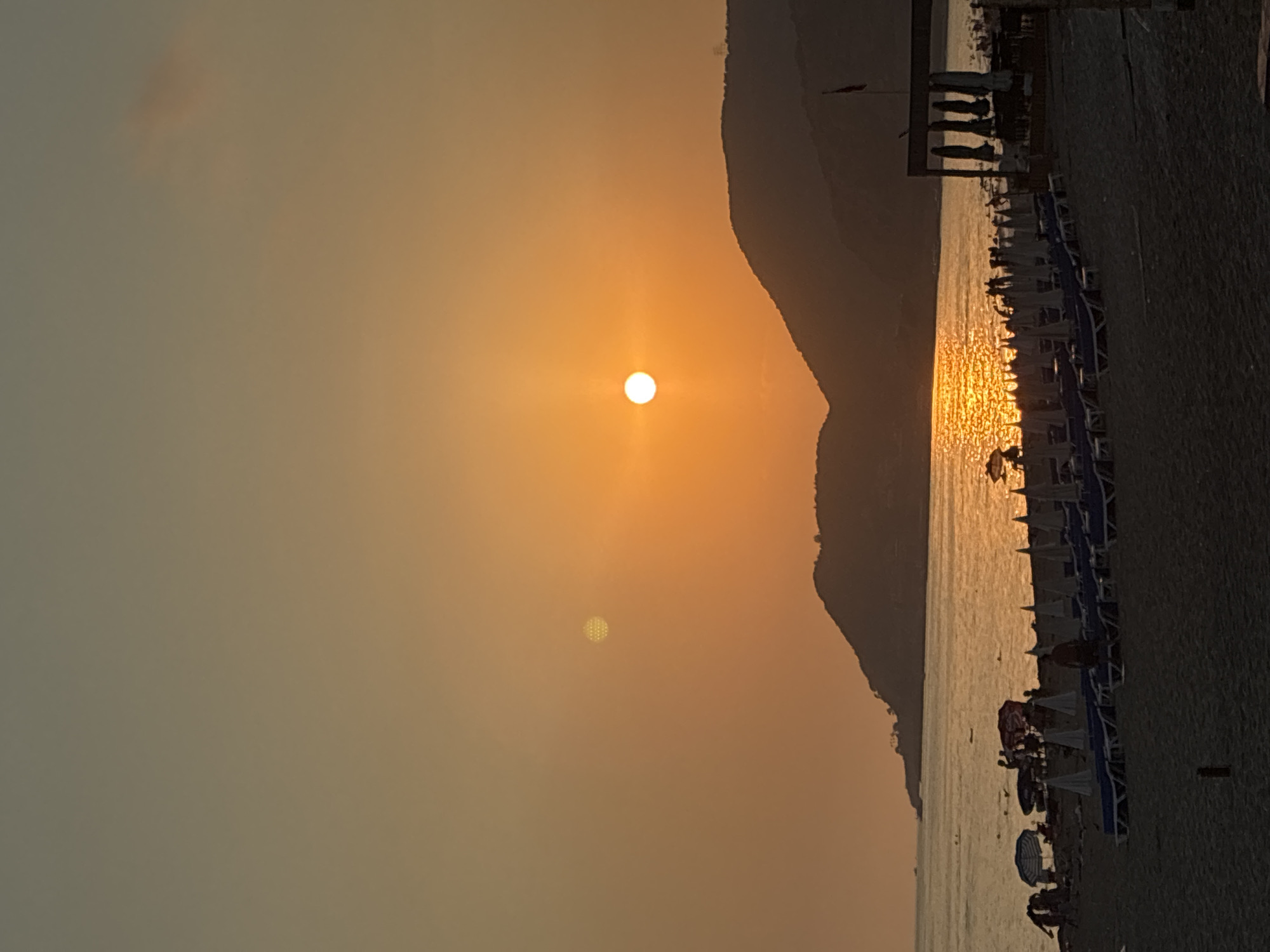 This is a vertical, eye-level, medium shot of a beach at sunset in Alanya, Turkey. The sun is a bright orange sphere in a hazy orange sky, casting a golden sheen on the water. The foreground is a sandy beach with rows of empty lounge chairs and closed umbrellas. Further back, some people are visible near the water's edge, with more lounge chairs and umbrellas set up closer to the sea. A dark mountain range forms the horizon, silhouetted against the vibrant sky. The overall atmosphere is serene and tranquil.